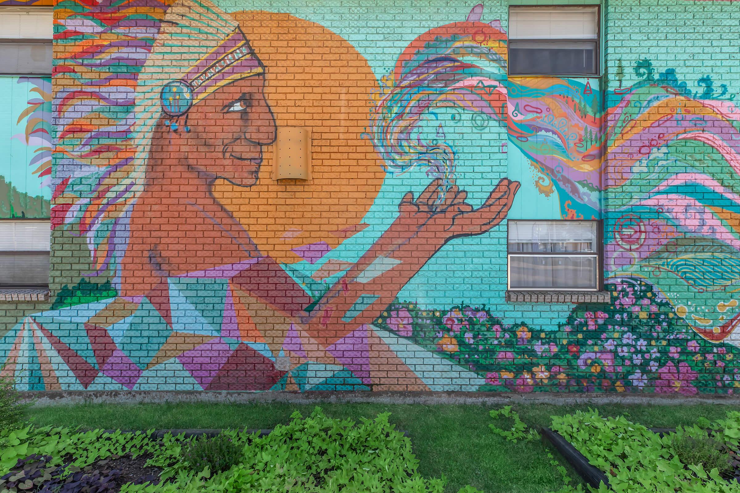 A colorful mural depicting a stylized figure, resembling a Native American, with vibrant patterns and a large sun behind. The figure is blowing colorful waves of energy or smoke, surrounded by floral designs and abstract shapes on a brick wall. The scene conveys a sense of creativity and connection to nature.