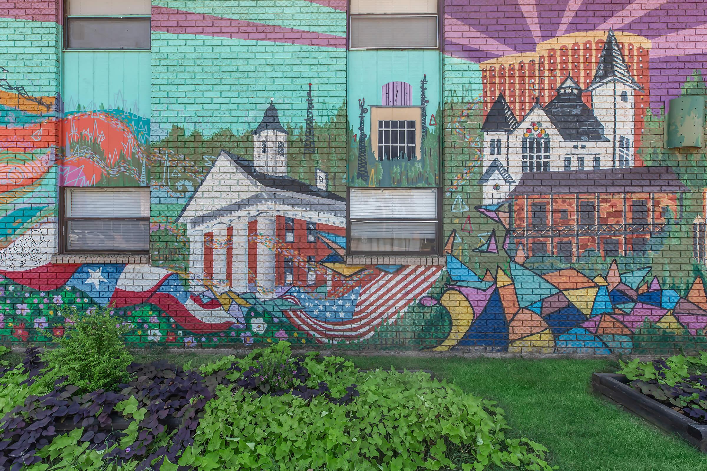 A vibrant mural depicting a colorful landscape with a mix of historical buildings, including a church and a courthouse. The artwork features elements like the American flag, lush greenery, and abstract shapes, all set against a bright background of blues, greens, and purples, showcasing local culture and heritage.