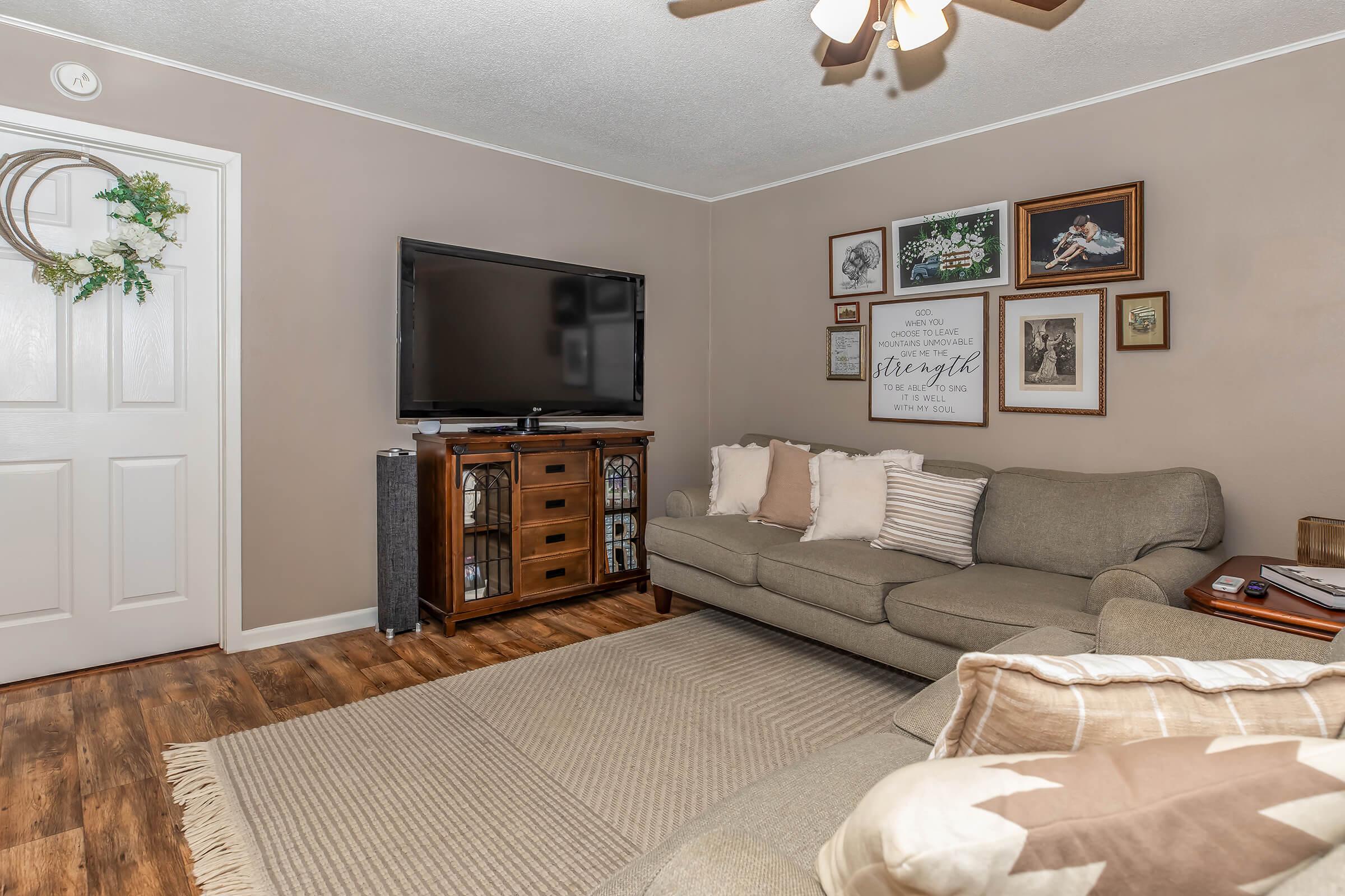 A cozy living room featuring a light brown sofa adorned with throw pillows. There's a wooden media cabinet beneath a flat-screen TV mounted on the wall. An area rug is on the floor, and a gallery wall of framed pictures and decor can be seen in the background, along with a decorative wreath on the door.