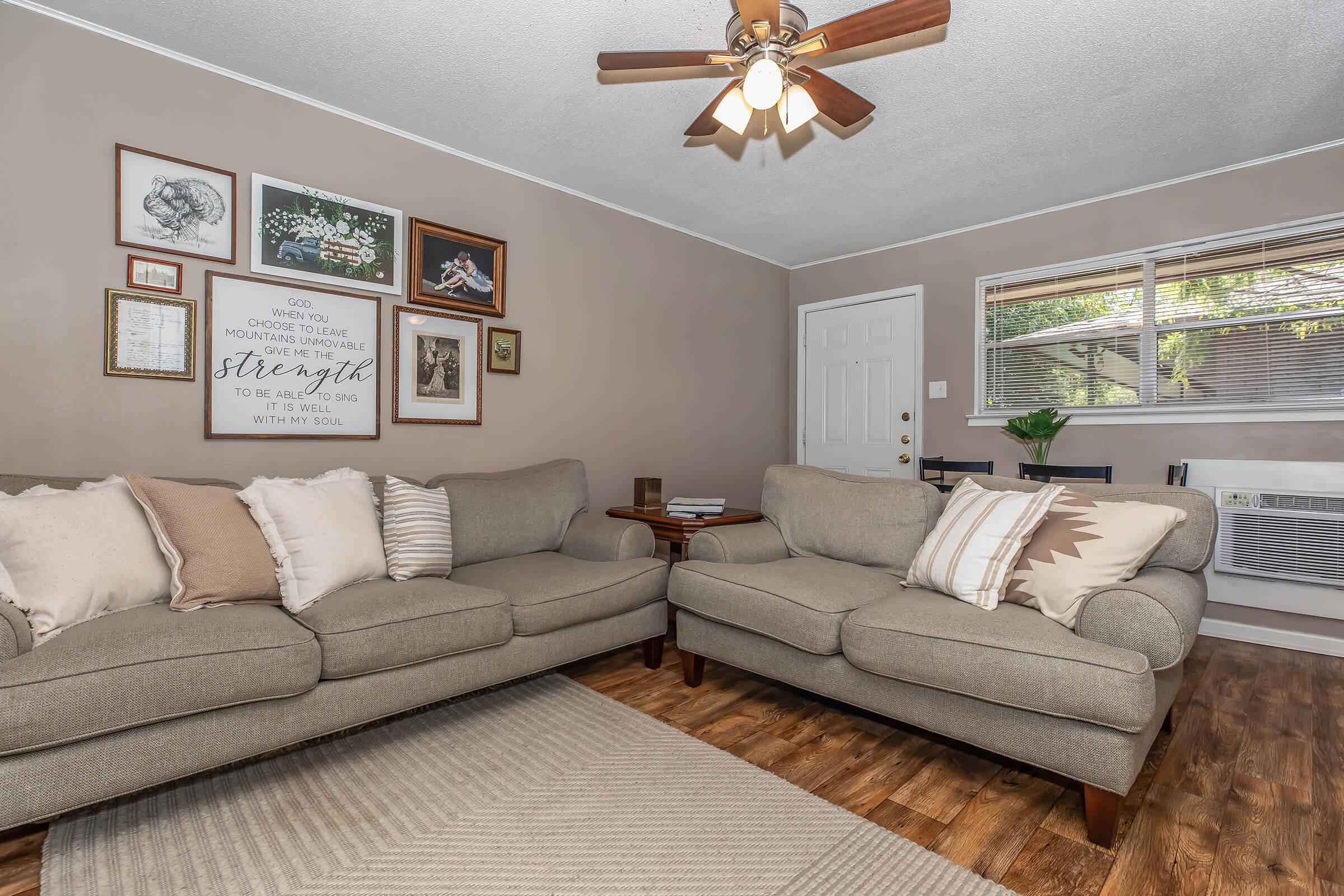 A cozy living room featuring two light gray sofas adorned with decorative pillows. The walls are decorated with framed artwork and a motivational quote. A wooden side table sits between the sofas, and there's a small dining table visible in the background. The room has a warm, welcoming atmosphere.