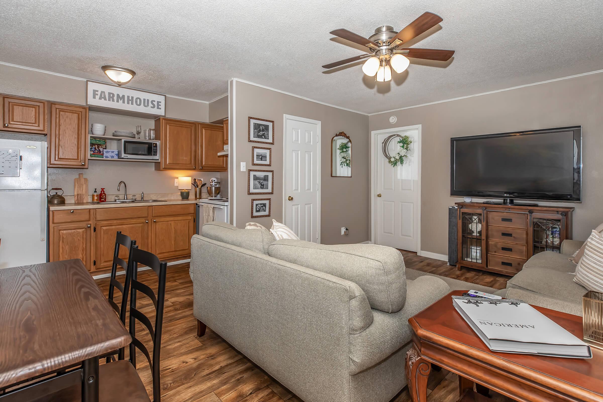 Cozy living room and kitchen area featuring a comfortable light gray sofa, wooden dining table with black chairs, and a flat-screen TV. The kitchen has wooden cabinets, and there are decorative elements like framed photos and greenery on the walls. A ceiling fan provides a homey atmosphere.