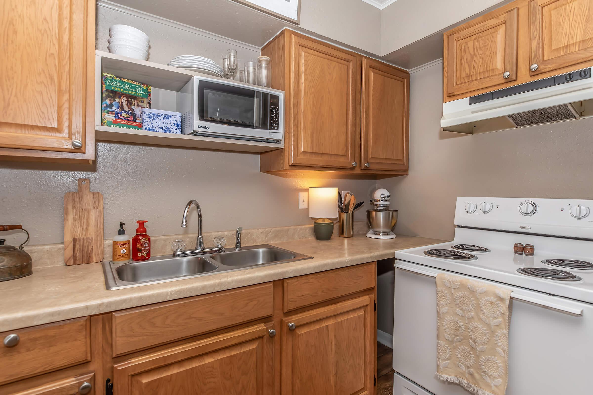 A modern kitchen featuring wooden cabinets, a double sink, a microwave, and a stove. The countertop is neatly organized with kitchenware, a lamp, and decorative items. Light-colored walls enhance the cozy ambiance, while natural light reflects off the surfaces, creating a warm atmosphere.