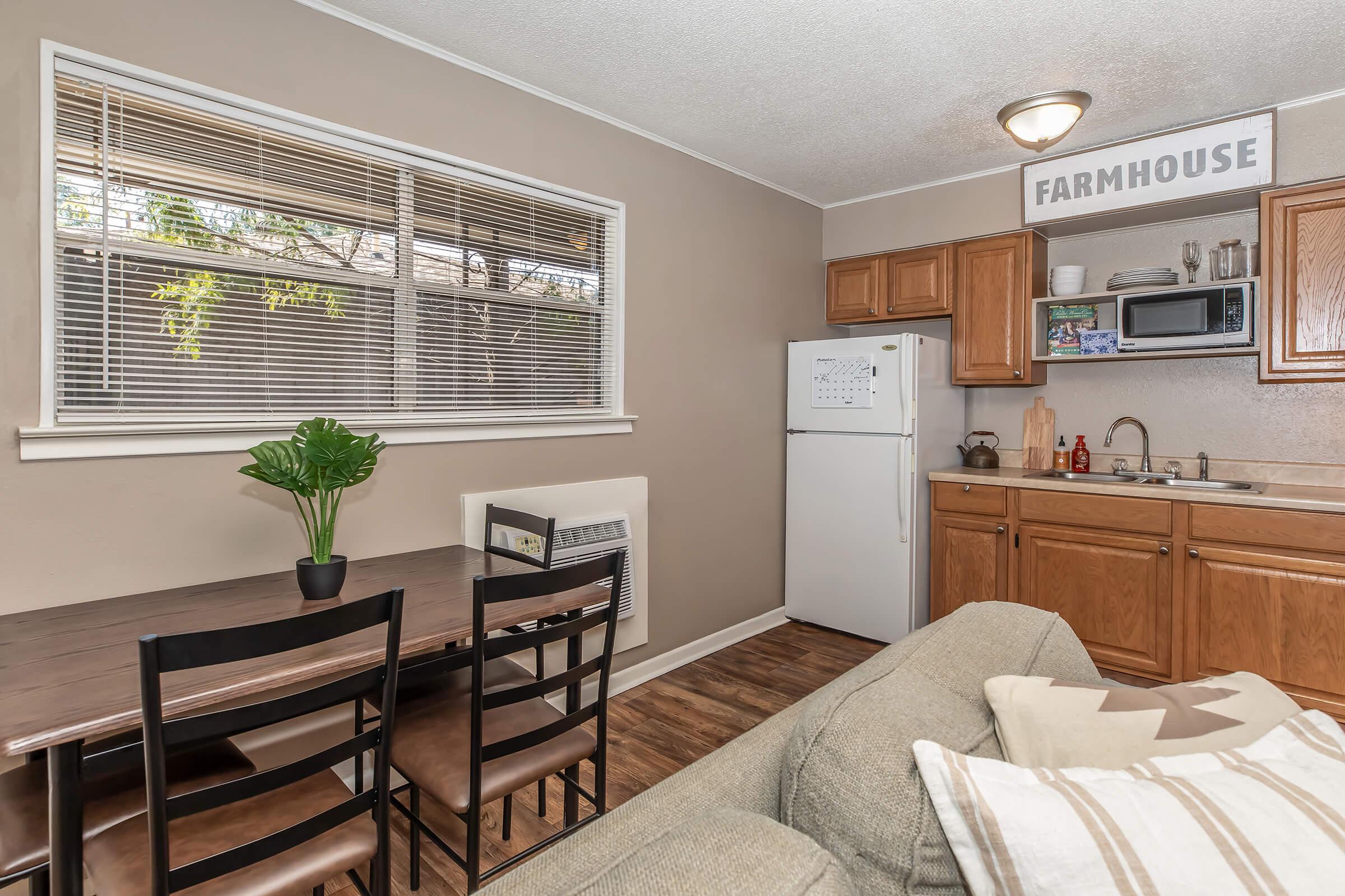 A cozy kitchen area featuring a wooden dining table with four chairs, a small refrigerator, and kitchen cabinets. There's a microwave and kitchenware on the counter. Natural light streams in through window blinds, and a potted plant adds a touch of greenery. The walls are painted in a neutral tone.