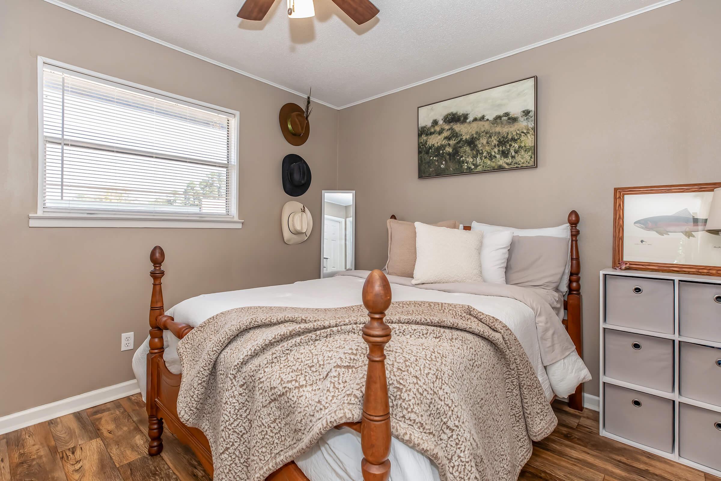 A cozy bedroom featuring a wooden bed with a light-colored blanket and decorative pillows. A small closet organizer sits by the bed, and hats are displayed on the wall above. A nature-themed art piece hangs above the bed, and a window provides natural light. The walls are painted a warm beige.