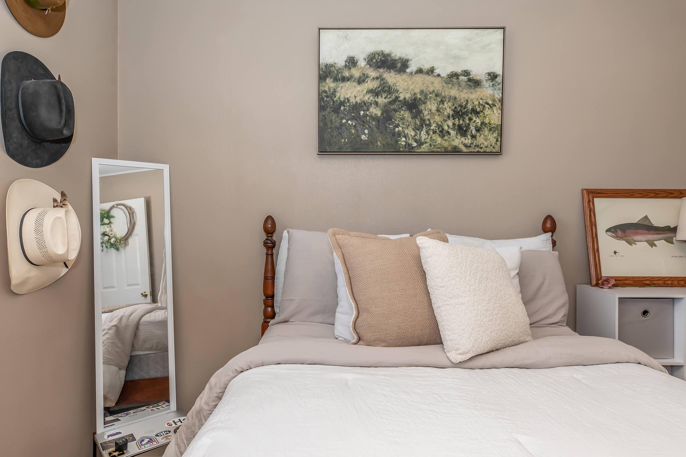 A cozy bedroom featuring a wooden bed with white and beige bedding, decorative pillows, and a mirrored wardrobe. The walls are painted in a warm tone, adorned with a landscape painting above the bed and a framed fish artwork nearby. Cowboy hats are displayed on the wall, adding a rustic touch to the decor.