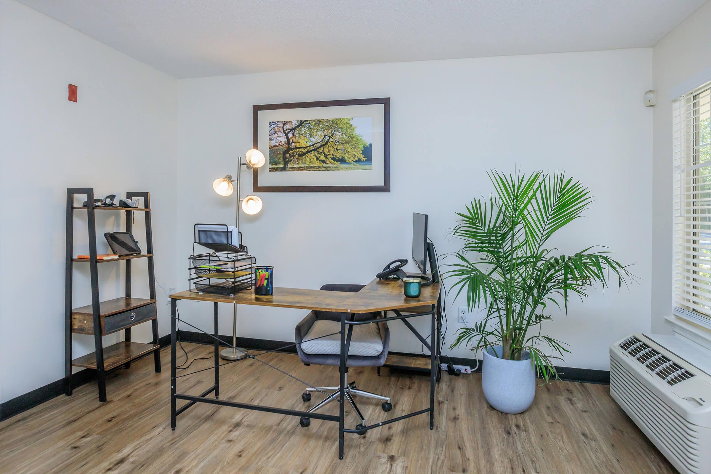 A modern home office featuring a wooden desk with a computer and a lamp, a black bookshelf with various items, and a large potted plant. There's a framed artwork on the wall and a window with blinds allowing natural light in. The floor is wooden, creating a warm atmosphere.