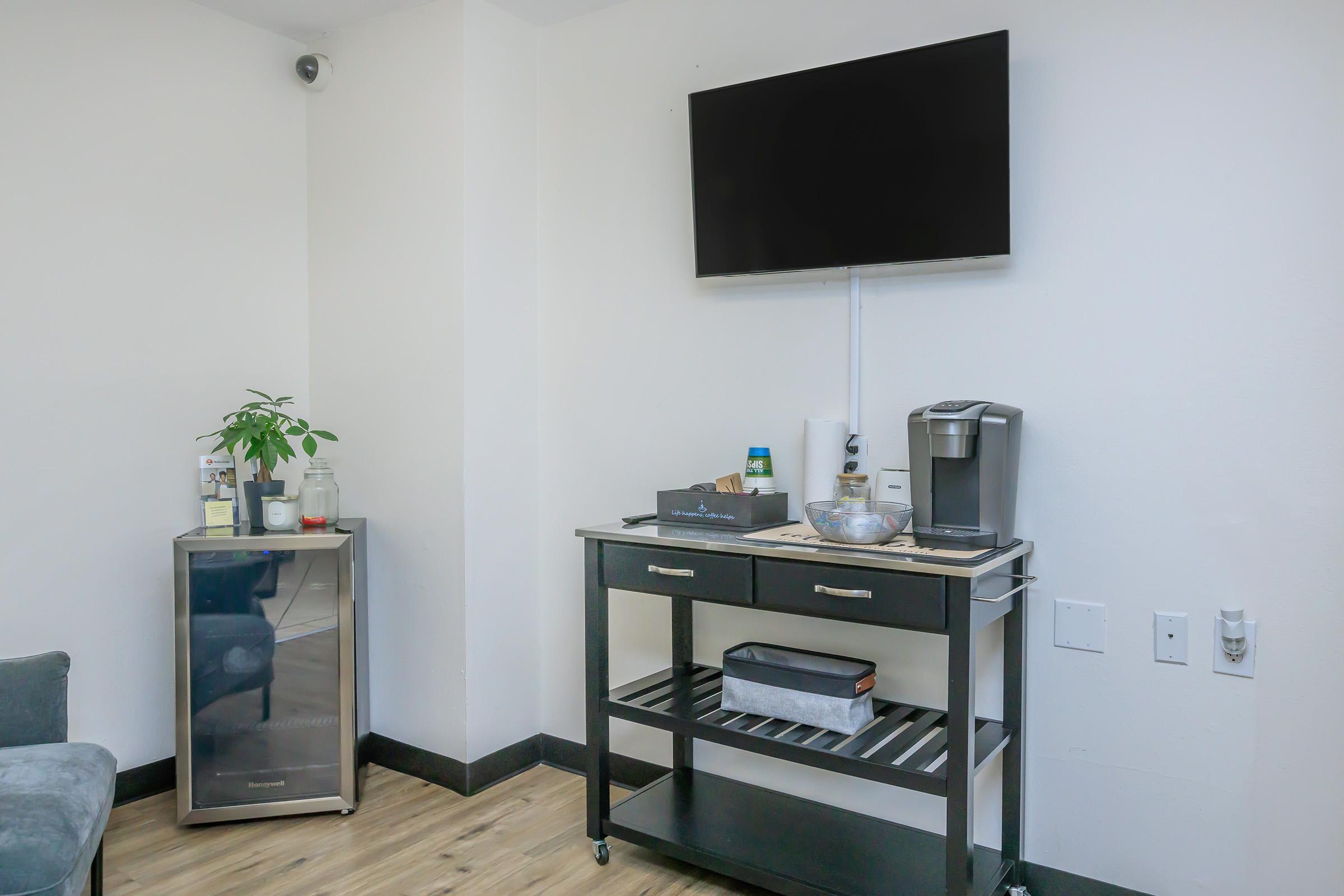 A small room featuring a wall-mounted TV, a coffee machine, and a plant on a cart. There is also a mini fridge next to the cart and a chair in the corner. The floor is wooden, and the walls are plain white.
