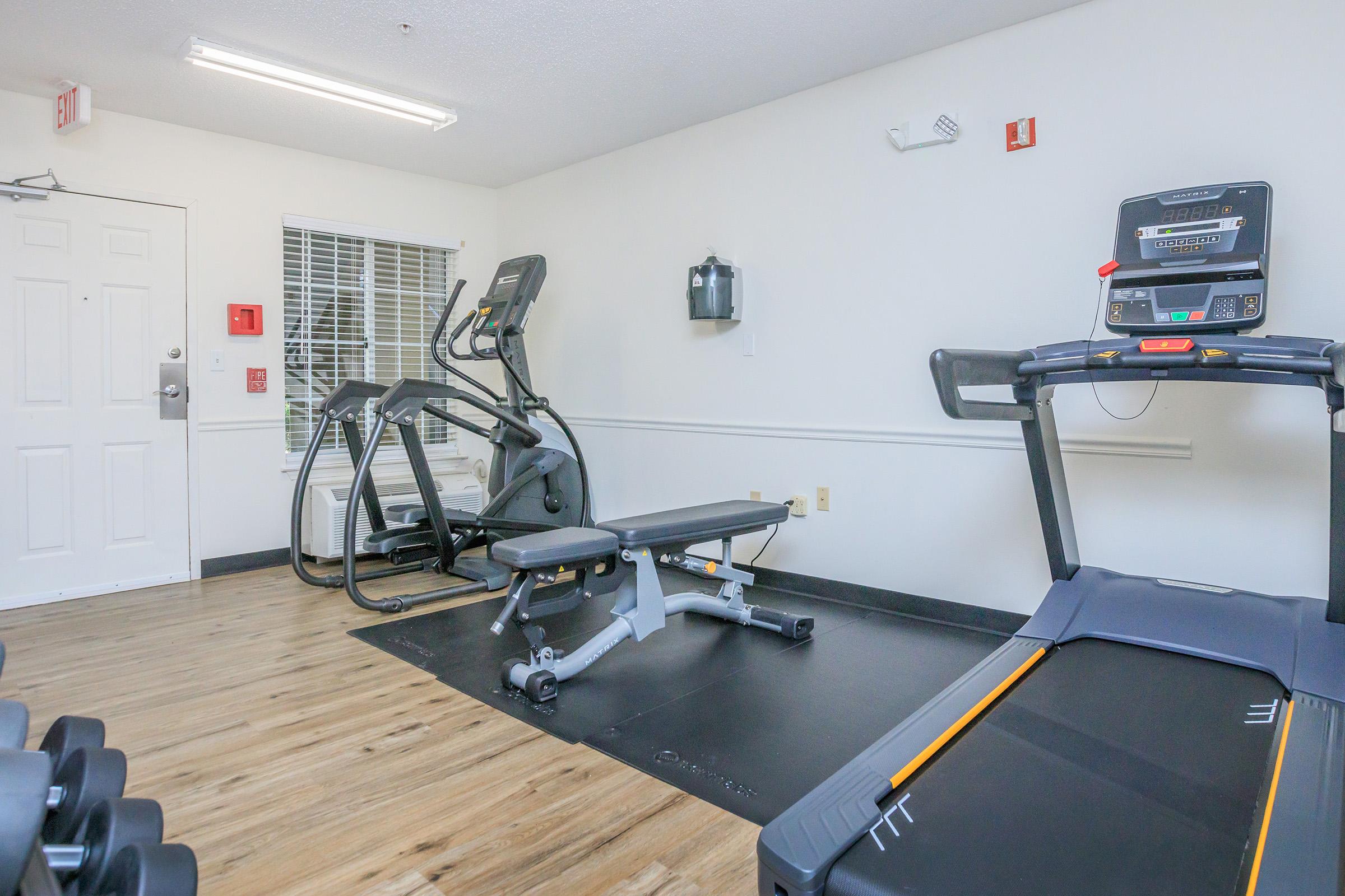 A fitness room featuring exercise equipment including a treadmill, elliptical machine, weights on a rack, and a workout bench. The walls are painted white, and there is a window letting in natural light. The floor is wood-look laminate, creating a bright and inviting space for workouts.