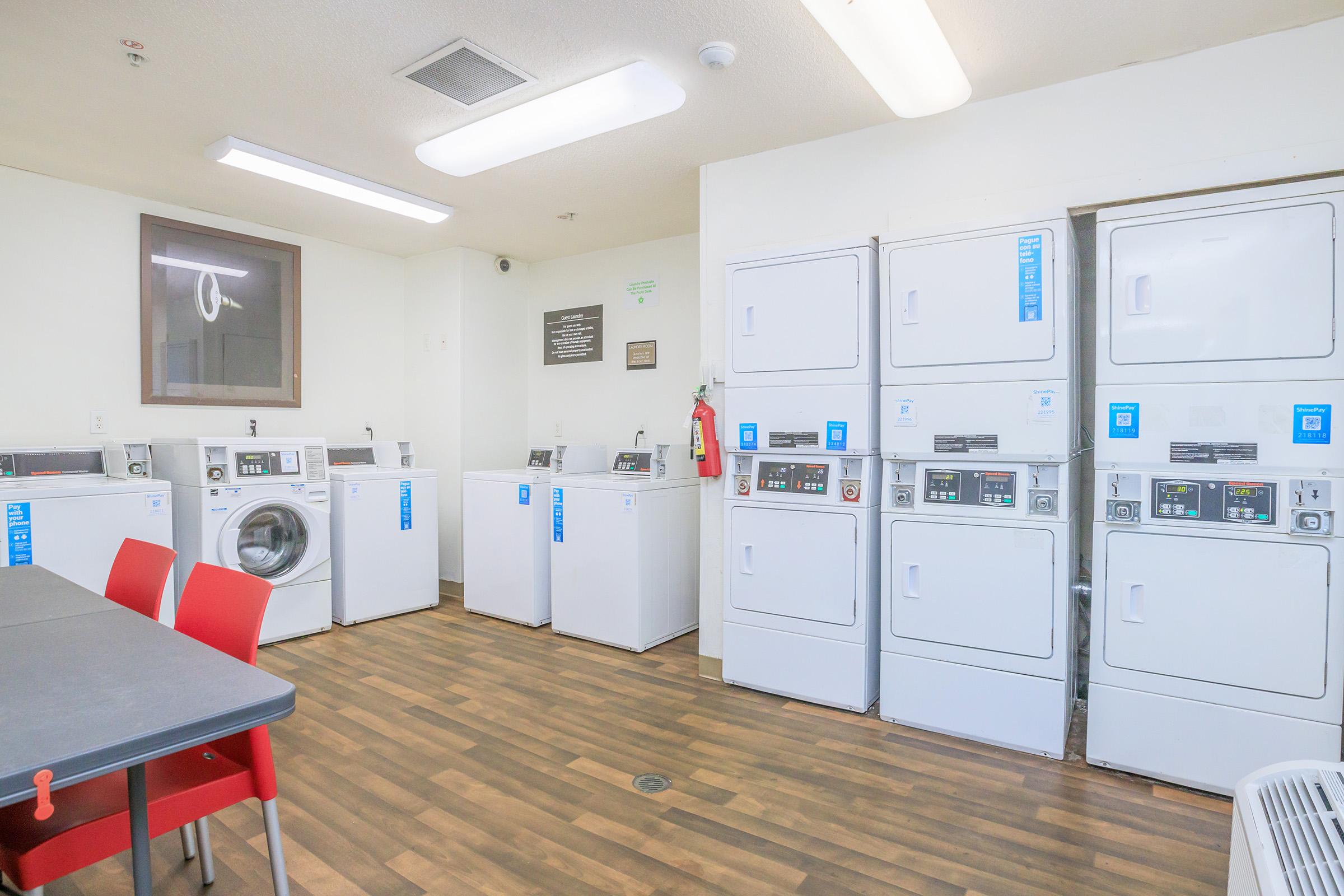 A clean laundry room featuring several washing machines and dryers arranged in a row. The machines are front-loading and have digital displays. There is a table with red chairs, and the flooring is wood-like. The walls are painted white, and there is a notice board and fire extinguisher visible.