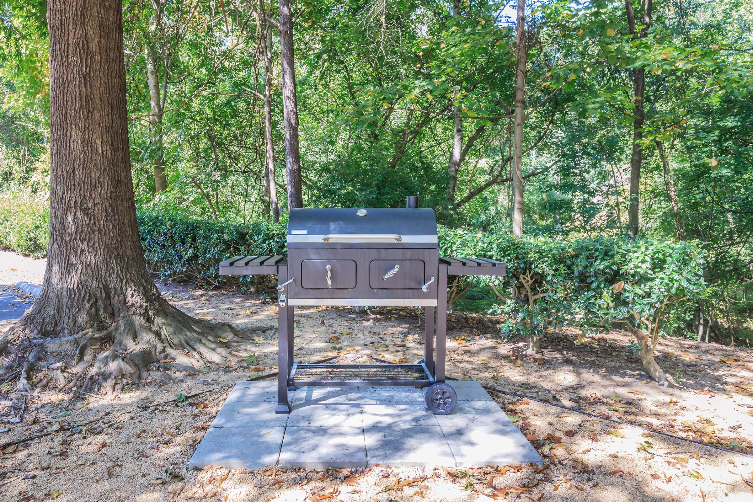 A black charcoal grill situated in a wooded area, surrounded by trees and bushes. The grill has a flat cooking surface, two doors for accessing the cooking chamber, and wheels for mobility. It is placed on a stone pad on a patch of dirt among foliage.