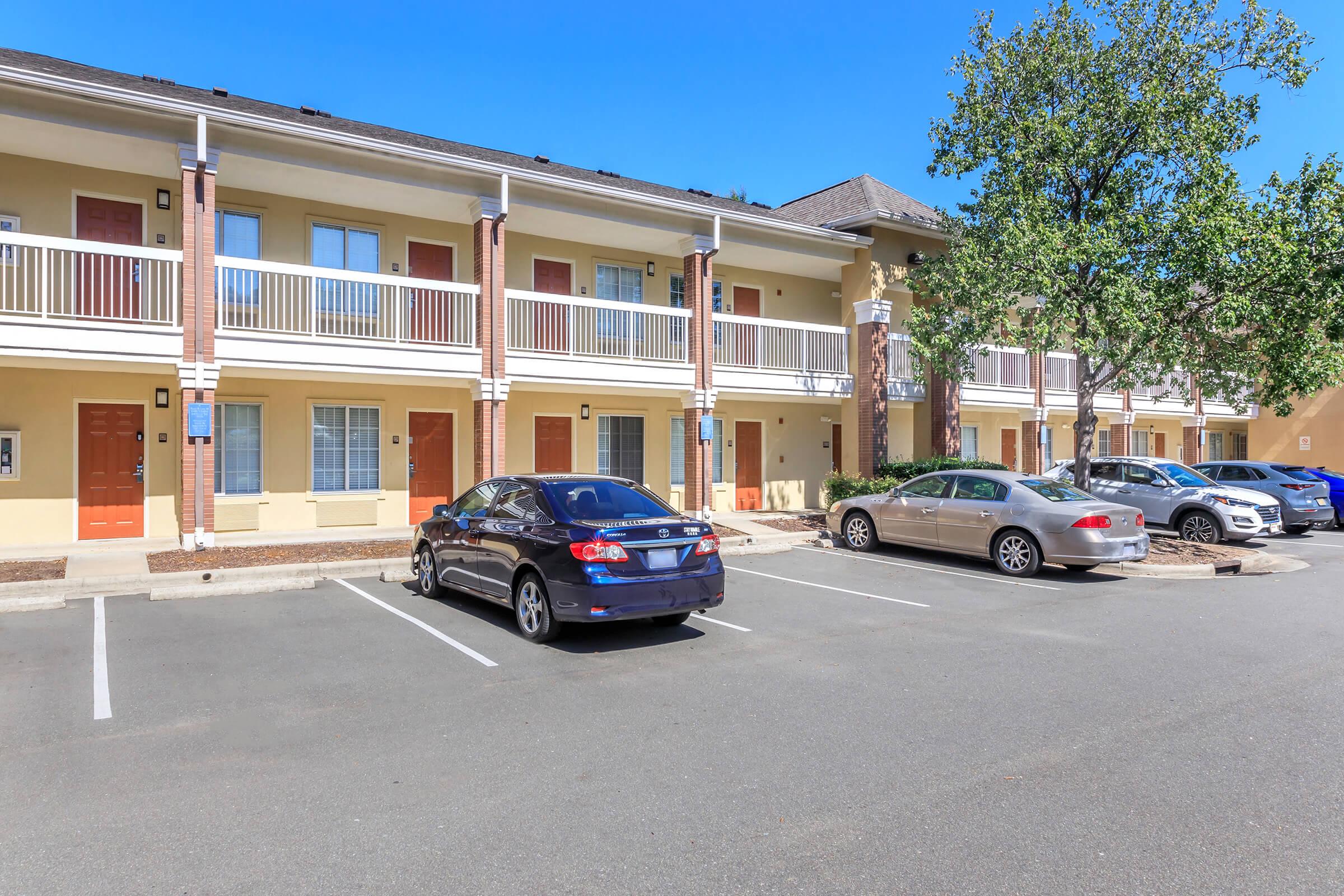 A view of a multi-story motel with several parked cars in the lot. The building features balconies, orange doors, and windows. The surrounding area is clear with a blue sky overhead, suggesting a sunny day.