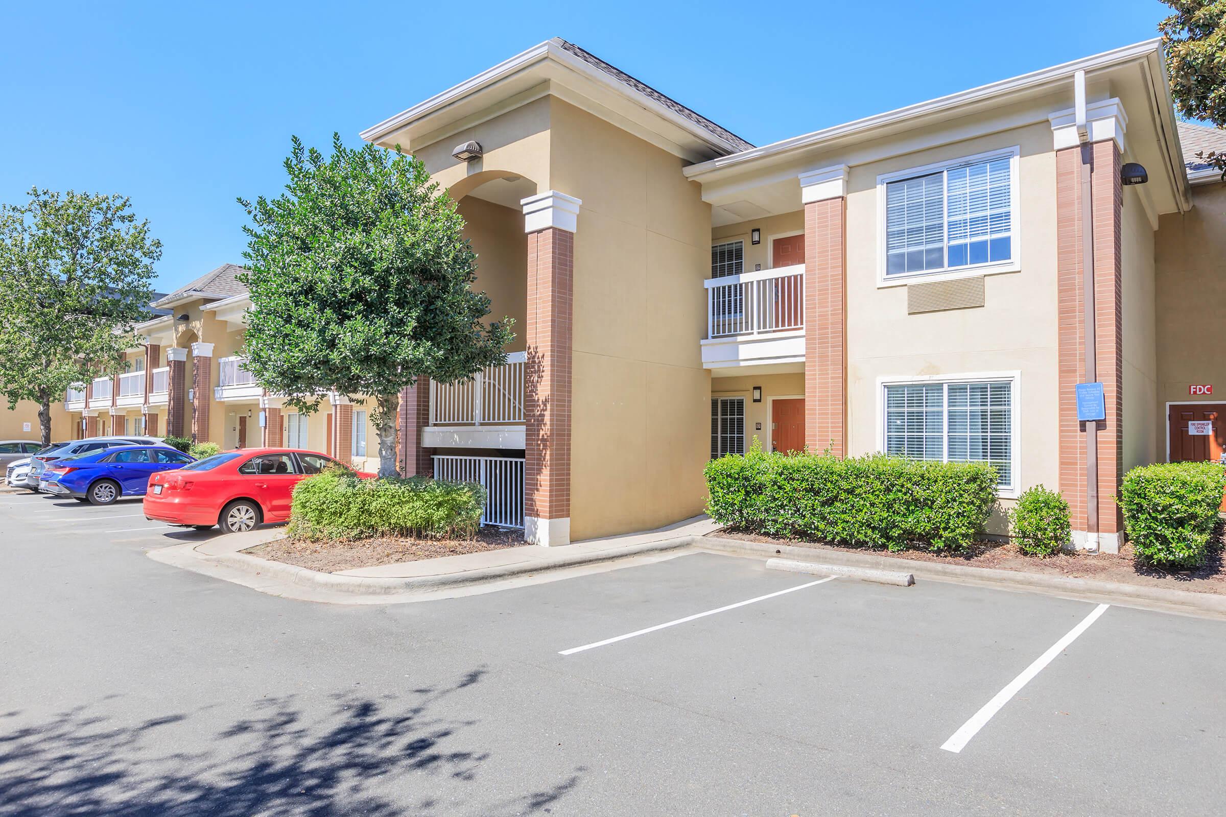 A well-maintained two-story apartment building with beige stucco walls and brick accents. The building features balconies, surrounded by neatly trimmed shrubs and trees. There are several parked cars, including a red sedan, in a spacious parking lot under a clear blue sky.