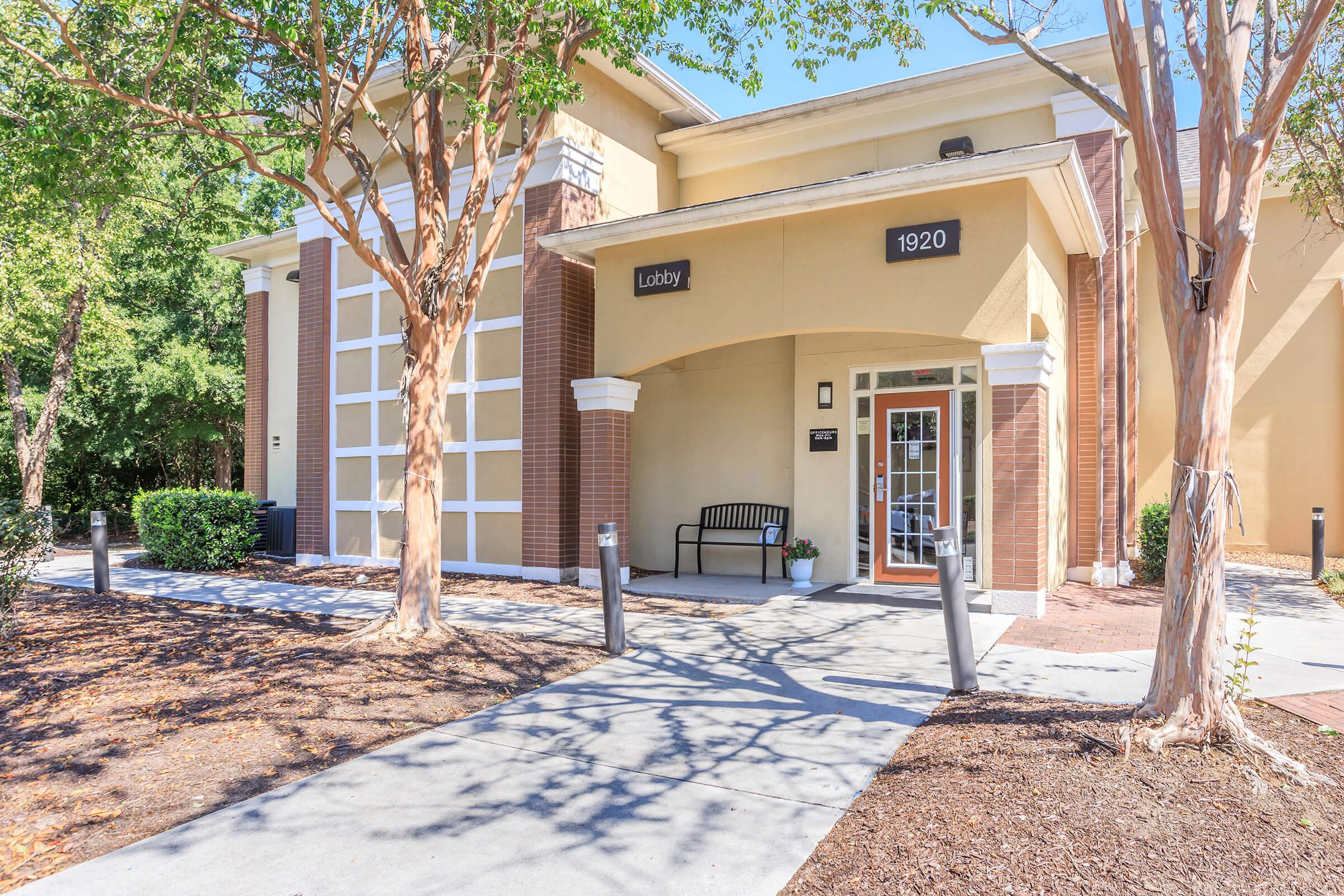 Exterior view of a modern building labeled "Lobby" with a welcoming entrance. A bench is positioned on the porch under a roof, surrounded by trees. The pathway leads up to the door, with landscaped areas on either side. Sunlight casts shadows on the ground, creating an inviting atmosphere.