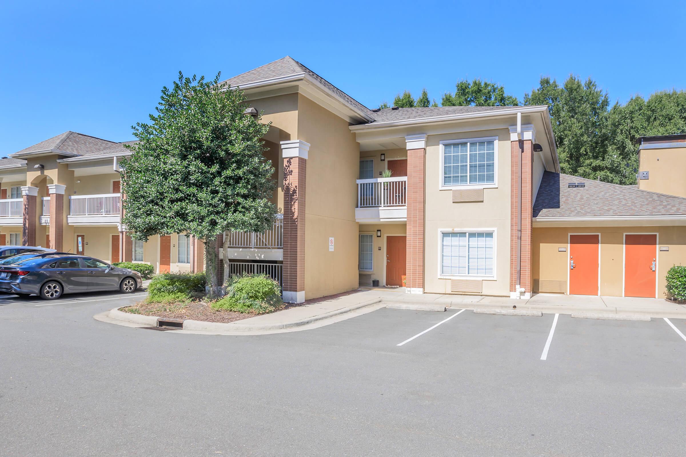 A well-maintained two-story apartment building with a light beige exterior, featuring balconies on the upper level. The parking lot is nearly empty, with a few cars parked nearby. Lush green trees and landscaping enhance the setting against a clear blue sky.