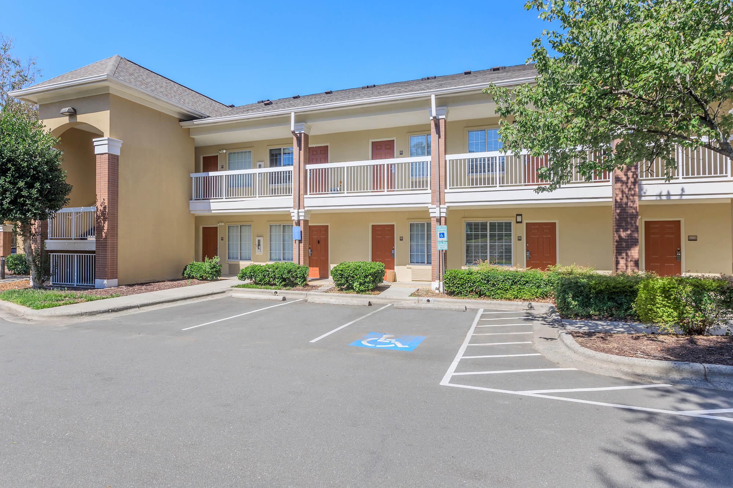 A view of a two-story building with multiple hotel rooms. The exterior features balconies and doors in various shades. In the foreground, there is a parking lot with a designated accessible parking space. The landscape includes small shrubs and trees, under a clear blue sky.