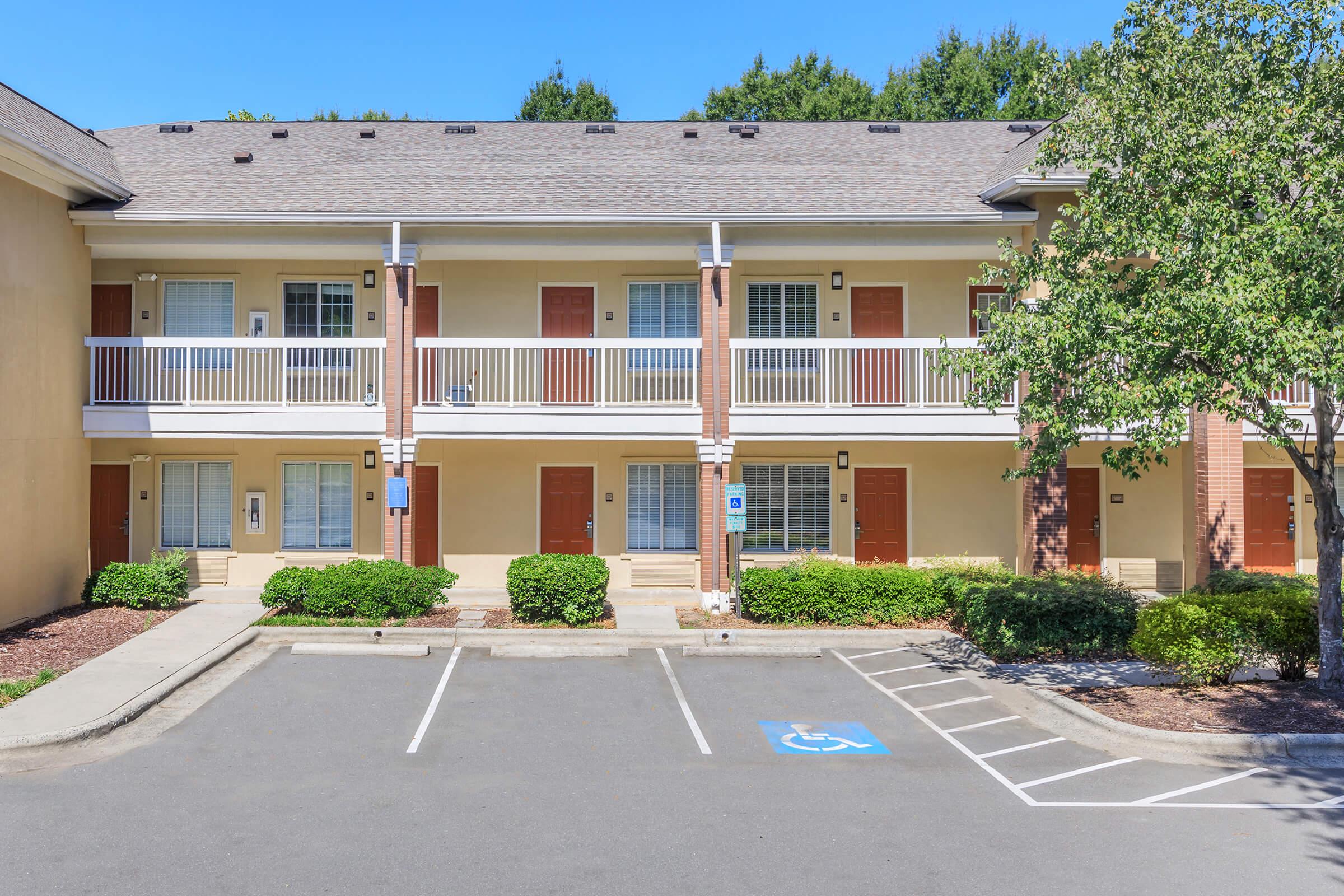 A two-story motel-style building with a light yellow exterior and brick accents. Each unit has a small balcony with white railings, and several doors are visible. The parking area in front has designated spaces, including one for handicapped accessibility, surrounded by landscaped bushes and trees.