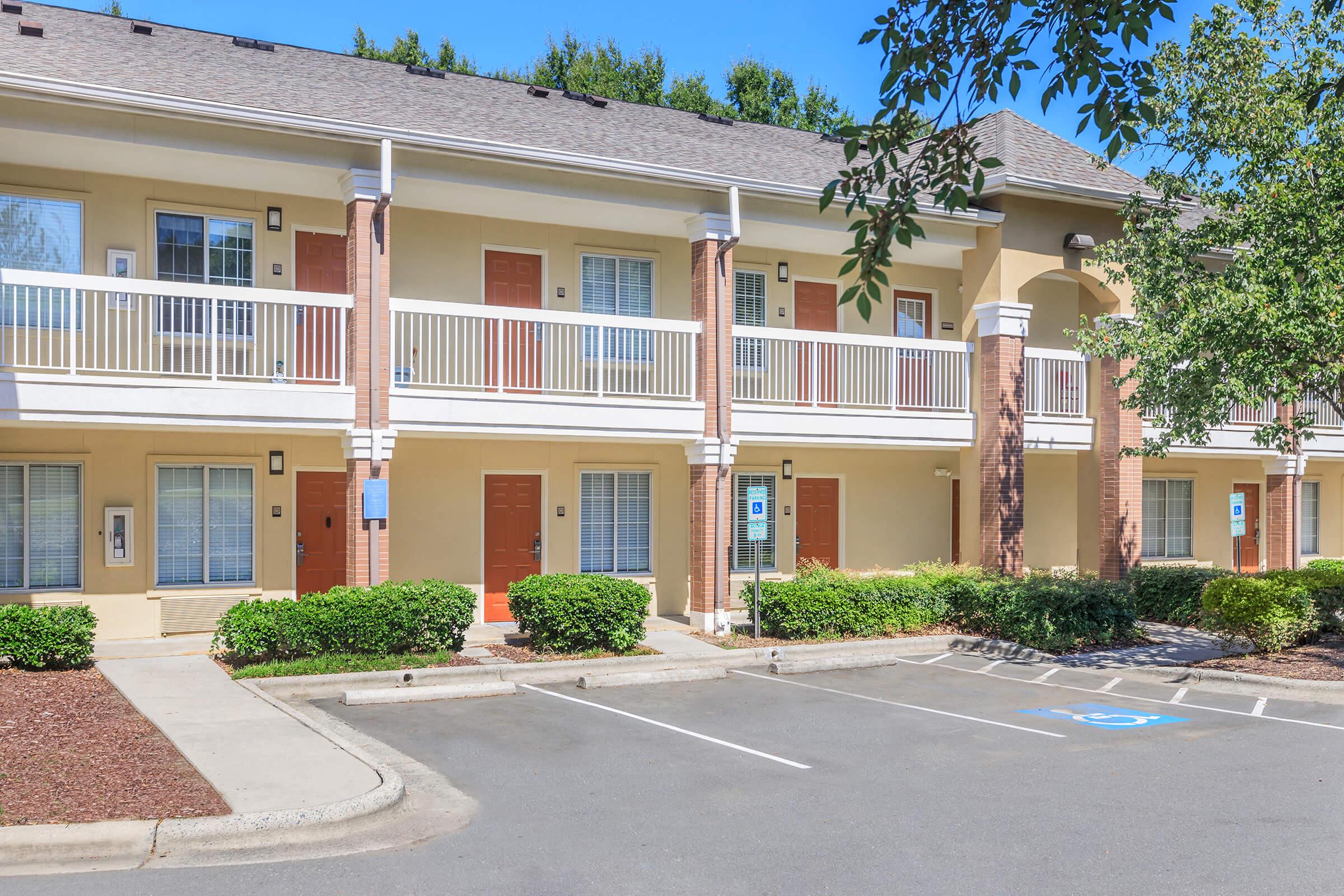 Exterior view of a two-story motel with beige siding and red doors. The building features balconies with white railings, surrounded by landscaped bushes. There is a parking lot in front, with a designated accessible parking space visible. A clear blue sky serves as the backdrop.