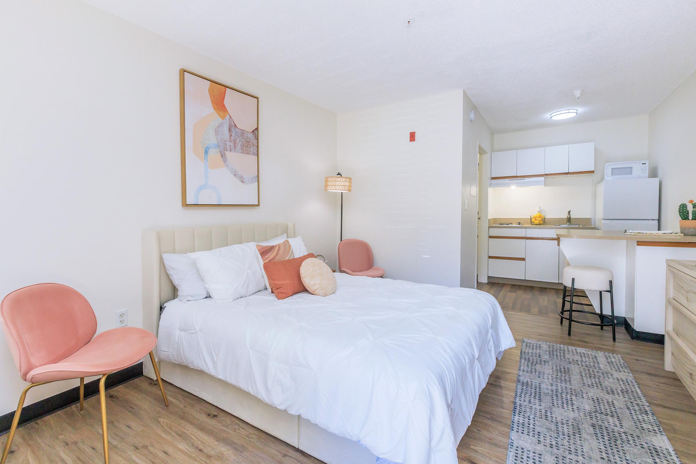 A cozy bedroom with a queen-sized bed adorned with pillows, a small kitchenette in the background, and a soft area rug on the floor. A lamp and two pink chairs add a touch of style, while an abstract painting hangs on the wall, contributing to the warm and inviting atmosphere.