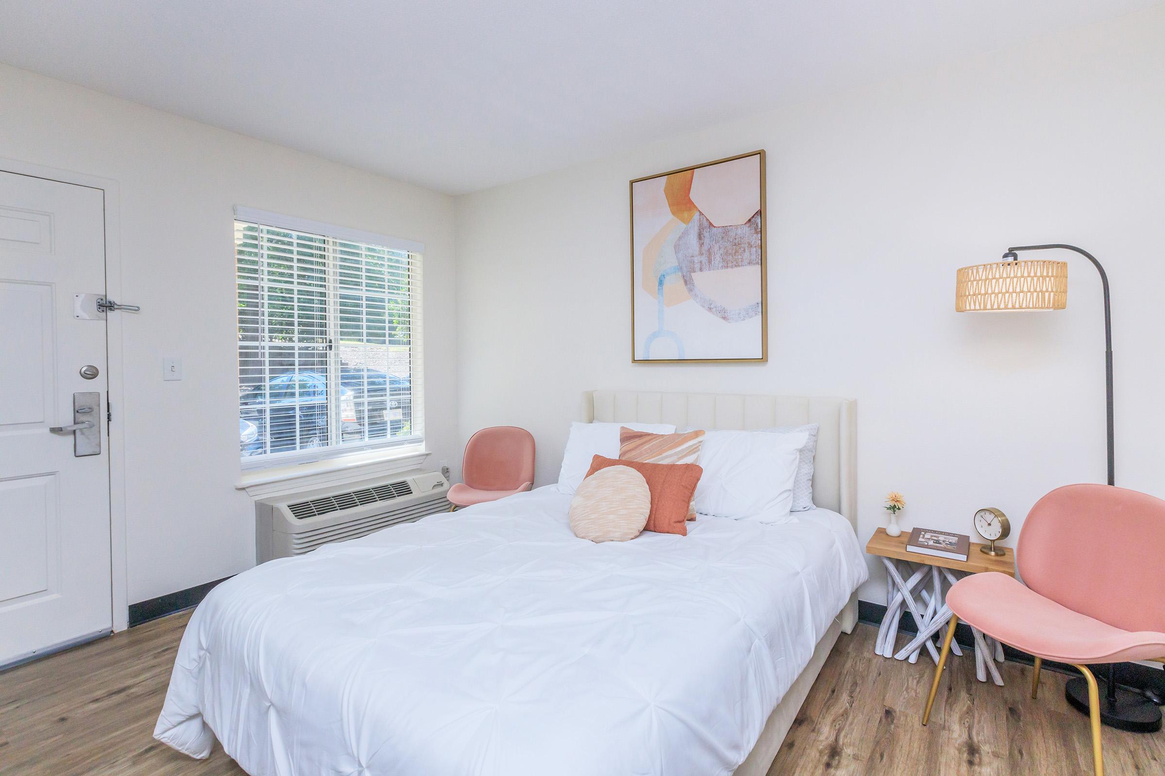 A cozy bedroom featuring a queen-sized bed with a white comforter, an orange decorative pillow, and natural wood flooring. There's a large window with sheer curtains, a bedside table with a clock, and a stylish lamp. Two pink accent chairs add a pop of color, while abstract artwork decorates the wall.