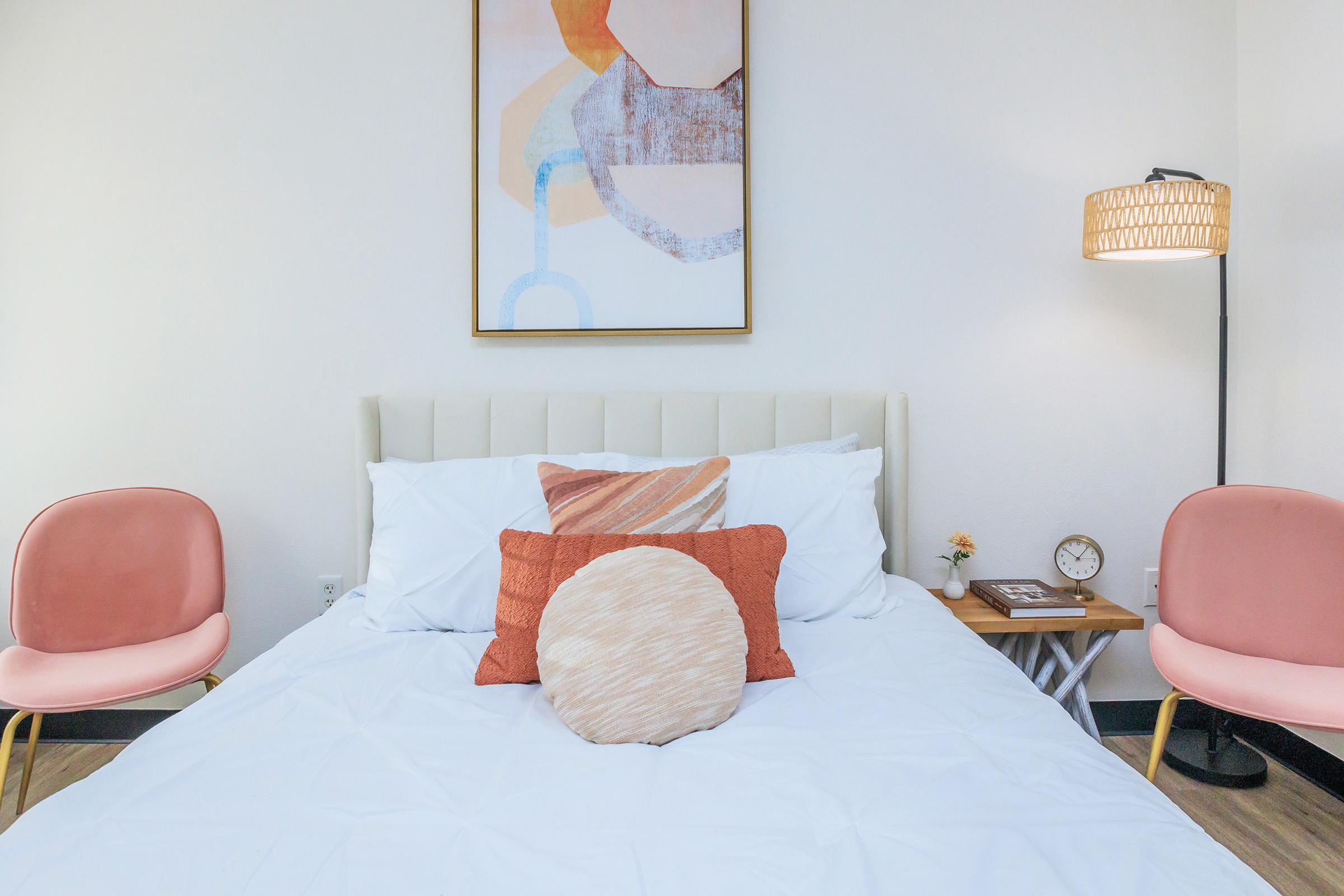 A cozy bedroom featuring a light-colored bed with decorative pillows, two pink chairs, a wooden side table with a small clock and a flower vase, and a stylish lamp. An abstract artwork hangs above the bed, adding a modern touch to the space. The overall ambiance is warm and inviting.
