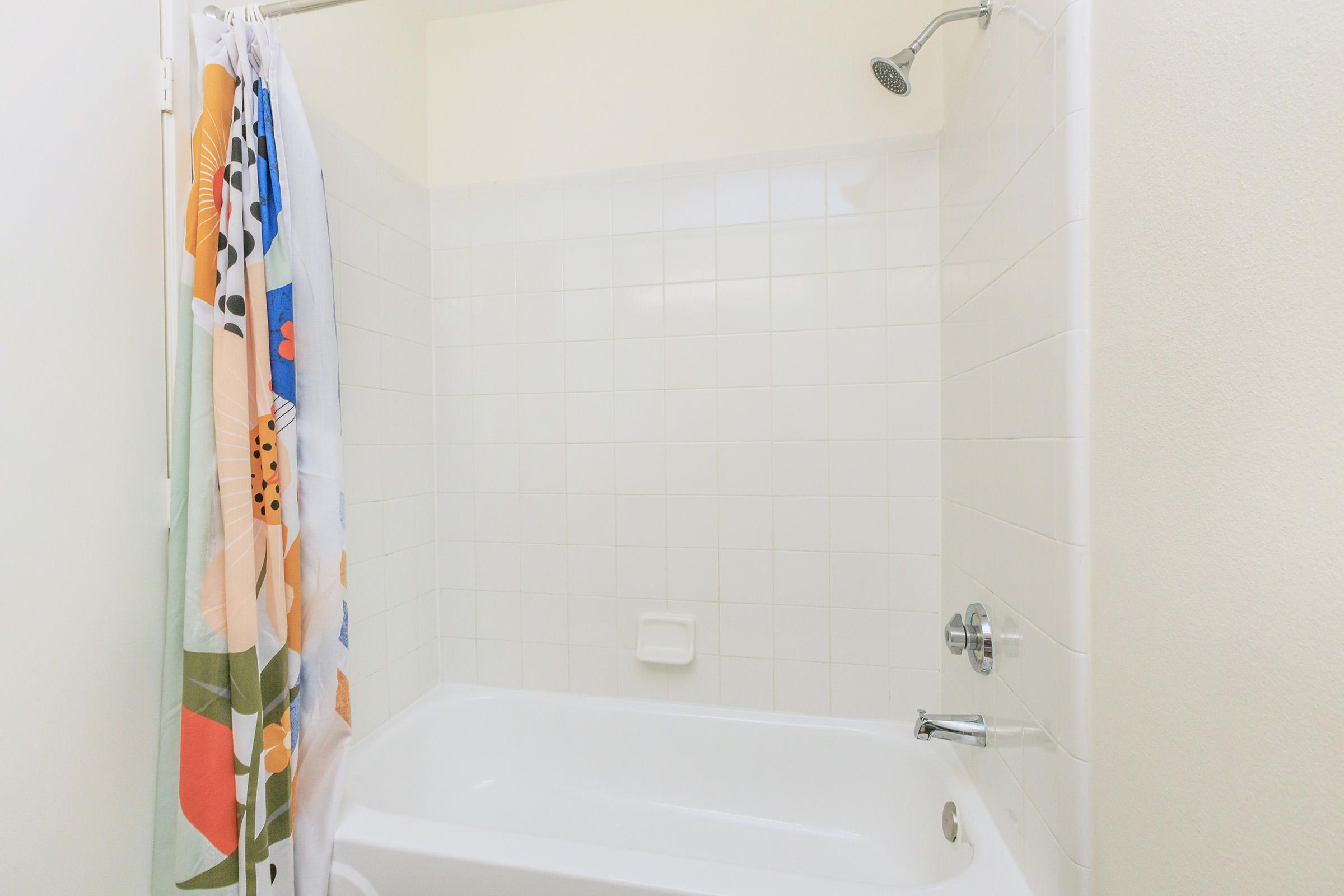 A clean bathroom featuring a white bathtub and a colorful shower curtain with floral and abstract designs. The showerhead is mounted on the wall, and white tiled walls surround the bathtub, creating a bright and airy feel.