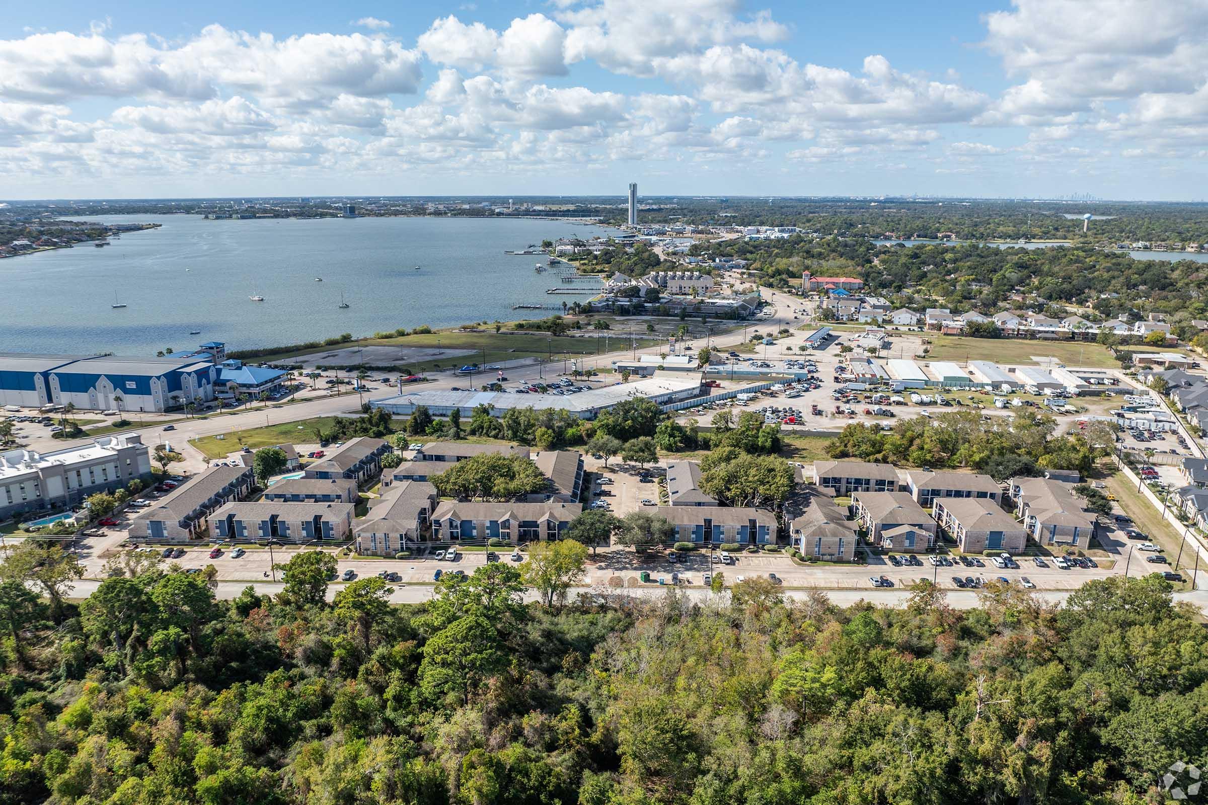 Aerial view of a coastal area featuring a mix of residential buildings and commercial spaces, with a waterfront in the background. Lush greenery borders the lower part of the image, and cars are parked on the streets. The sky is partly cloudy, creating a vibrant landscape.