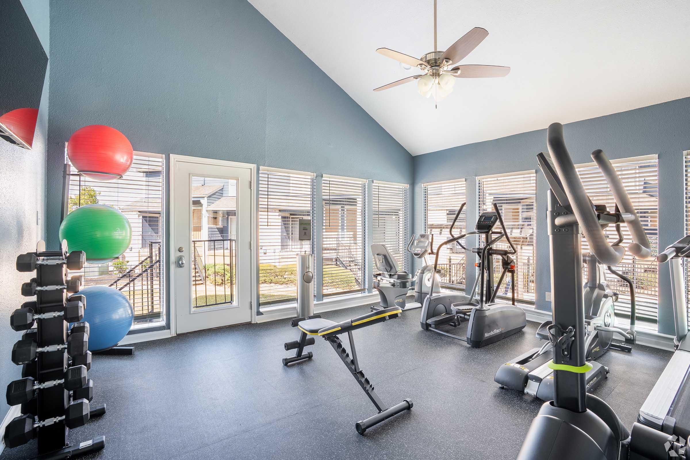 Bright and spacious gym area featuring exercise equipment like treadmills, stationary bikes, and weights. The room has large windows allowing natural light, with colorful exercise balls and a ceiling fan, creating an inviting atmosphere for workouts.