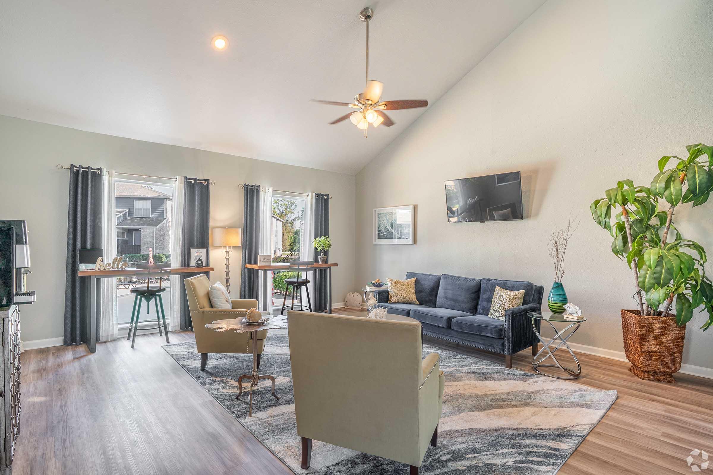 A spacious living room featuring a modern couch, two armchairs, and a coffee table. Bright windows with curtains provide natural light. A ceiling fan hangs above, and a television is mounted on the wall. Green plants and decorative items add warmth to the inviting space.