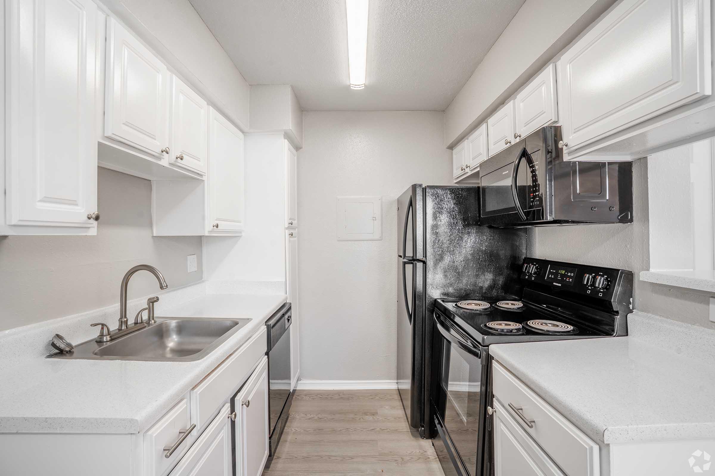 Modern kitchen featuring white cabinetry, a stainless steel sink, and black appliances including a microwave and electric stove. The countertop is light-colored with a seamless design, and the space has wood-style flooring, contributing to a clean and contemporary look.