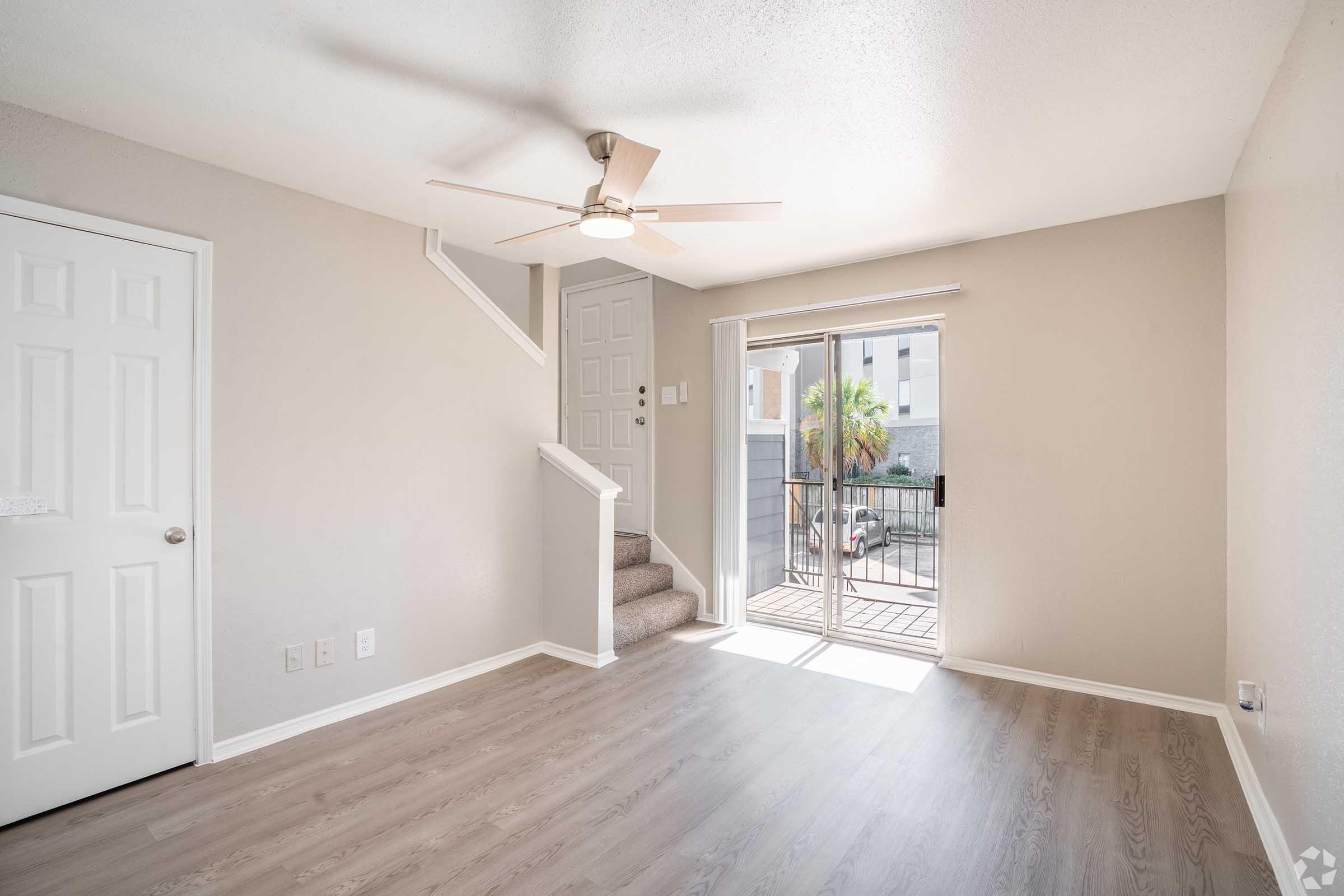 Interior view of a light and airy living room featuring a ceiling fan, a staircase leading to an upper level, and a sliding glass door that opens to a patio. The walls are painted a soft neutral color, and the floor has a wood-like finish. A closed door is visible on the left side.
