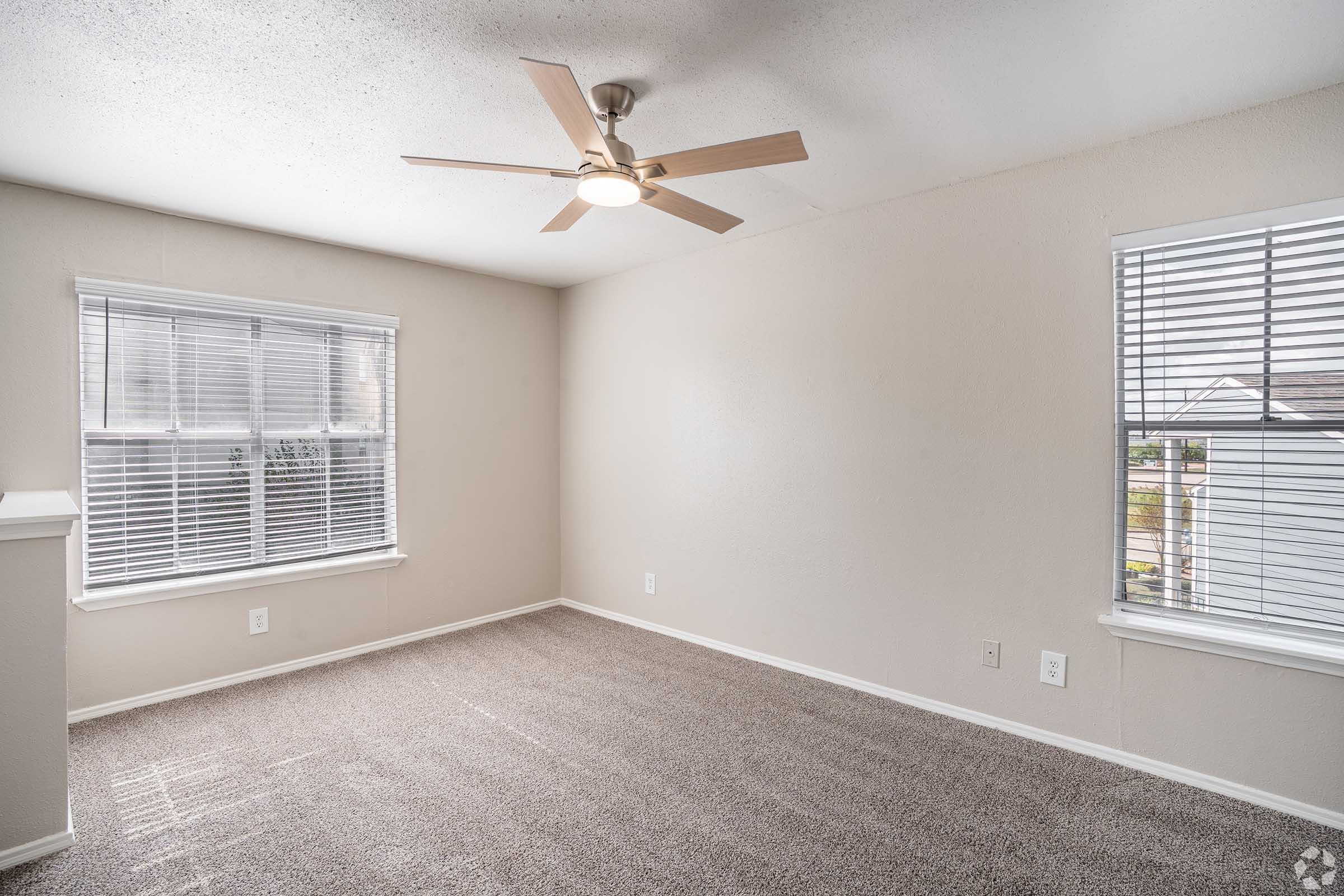 Empty room with beige walls and light brown carpet. Two windows with white blinds allow natural light to enter. A ceiling fan is mounted in the center of the room. The space appears clean and unfurnished, suitable for various uses.