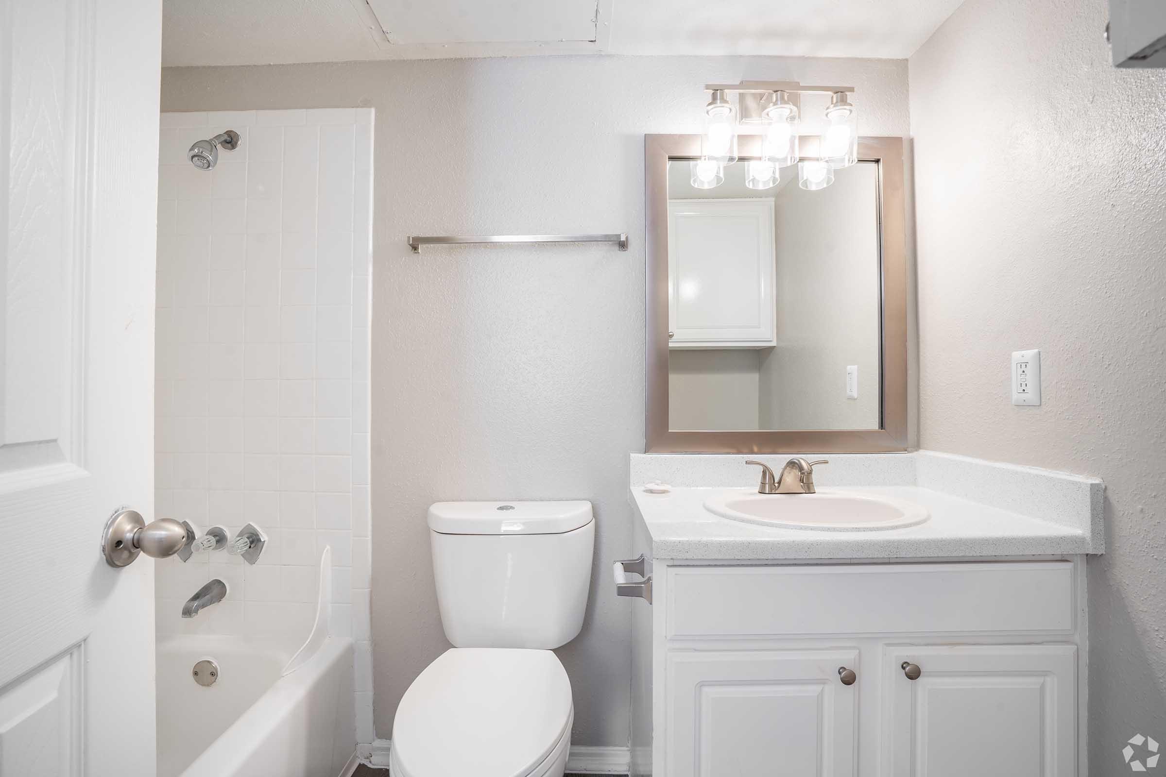 A clean, modern bathroom featuring a white bathtub and shower combo, a toilet, a single sink vanity with a mirror, and light fixtures. The walls are painted in a light gray color, and there are white cabinets for storage.