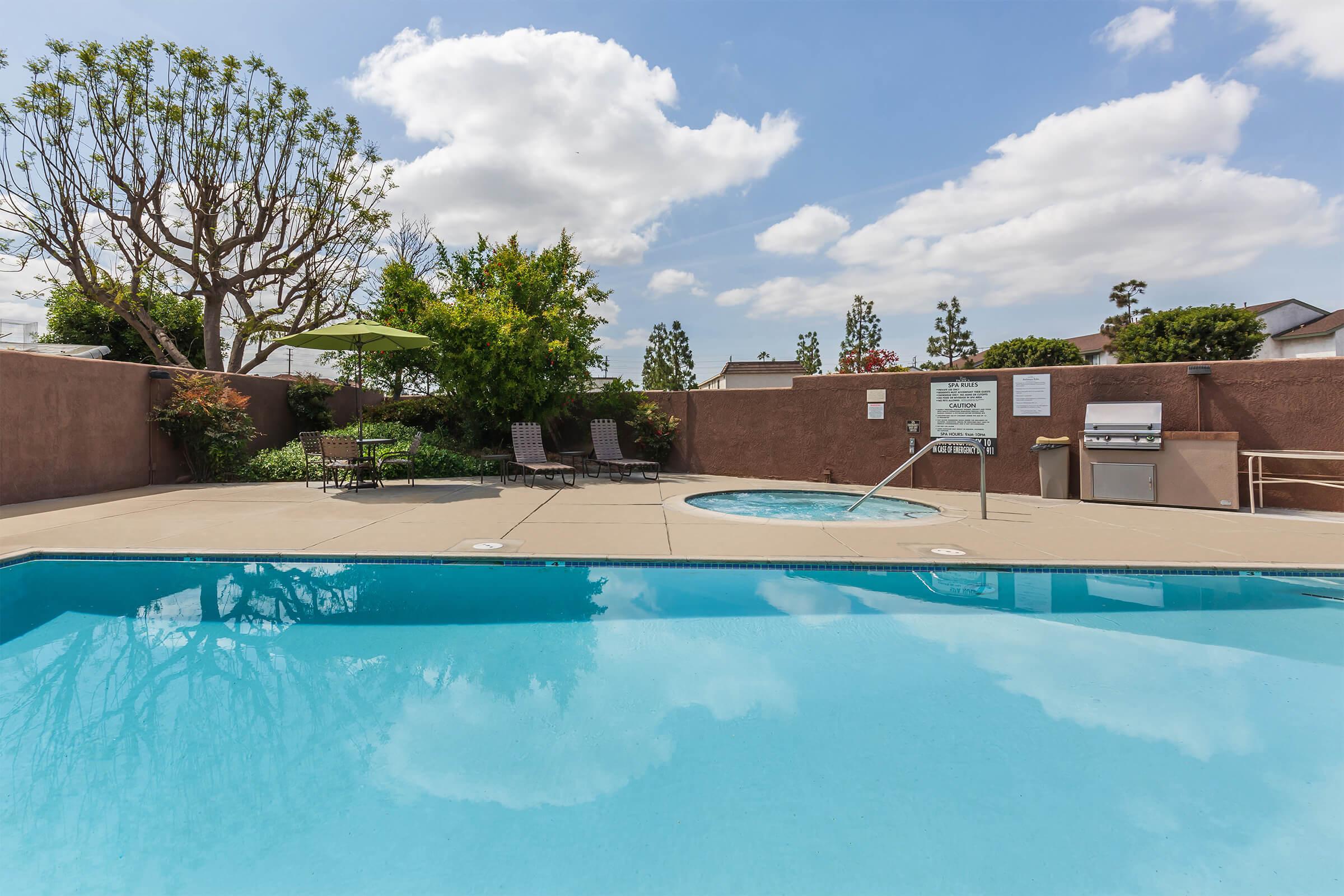 A sunny outdoor pool area featuring a clear blue swimming pool and a hot tub. Surrounding the pool are lounge chairs and a green umbrella, along with a barbecue grill nearby. Lush trees and shrubs provide a green backdrop against a partly cloudy sky.