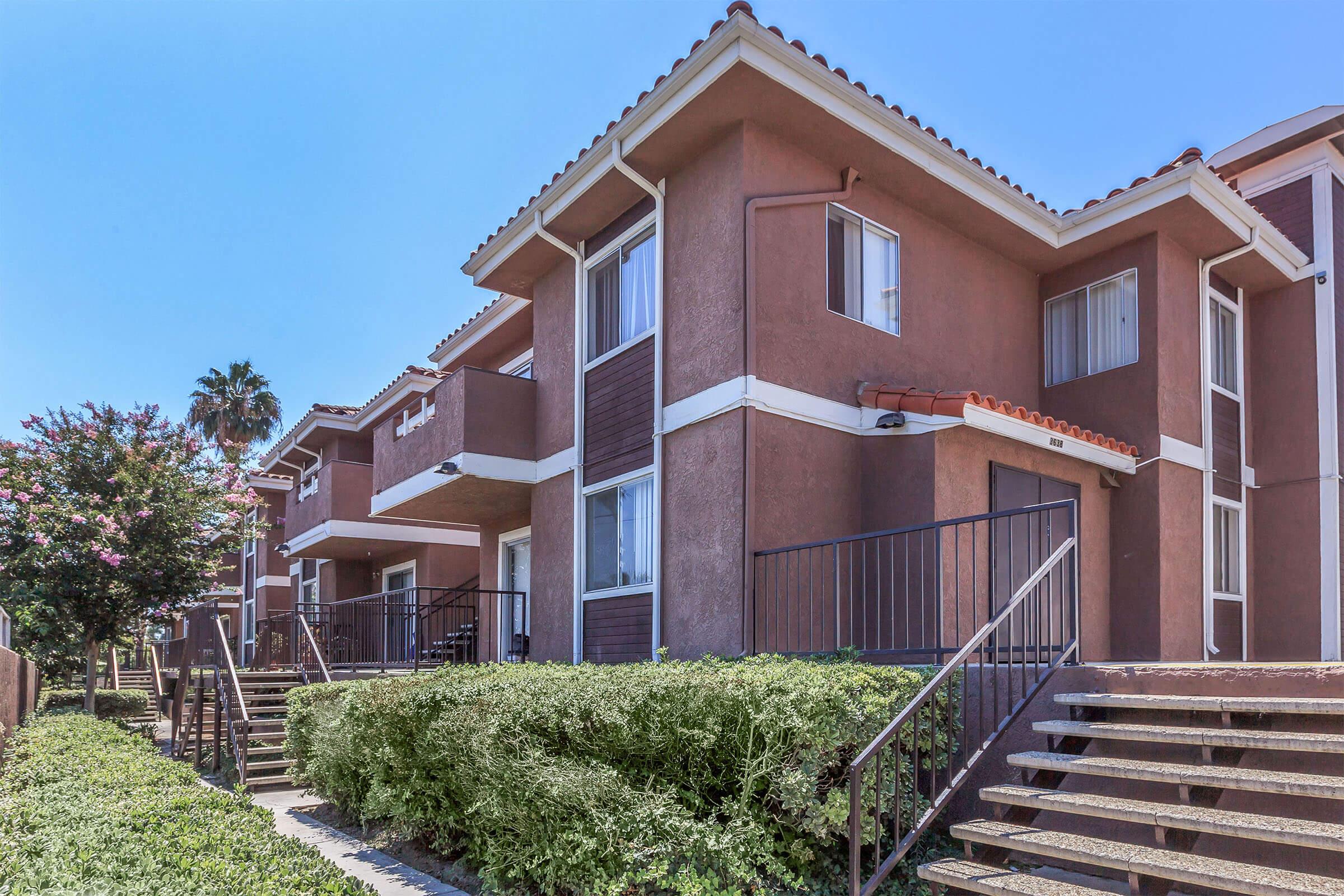 A view of a residential building with two-story apartments. The architecture features a mix of brown exterior walls and white trim, with balconies on the upper levels. There are landscaped shrubs and stairs leading up to the entrances, under a clear blue sky.