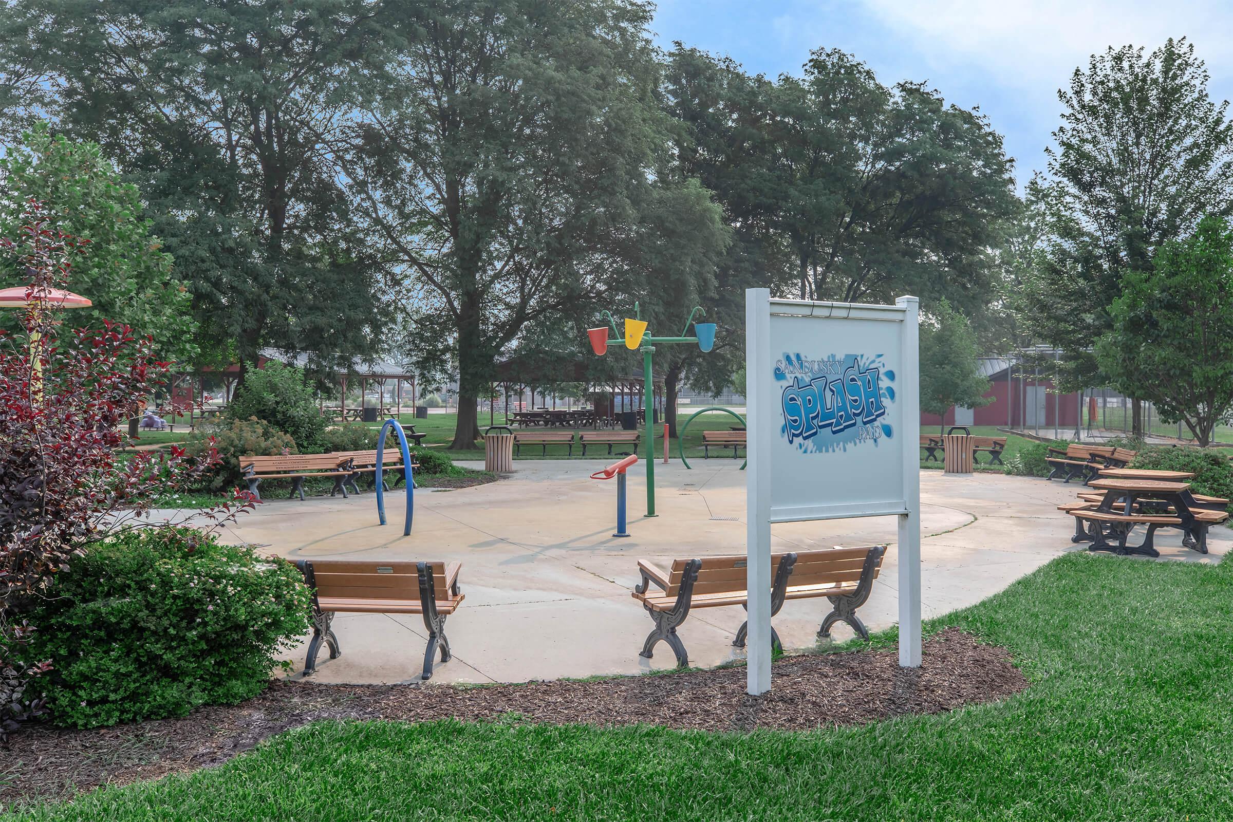 A vibrant splash pad area in a park featuring colorful water features, picnic benches, and lush greenery. The main sign reads "Splash," inviting visitors to enjoy the water play. Trees provide a scenic backdrop, enhancing the cheerful atmosphere of the space.