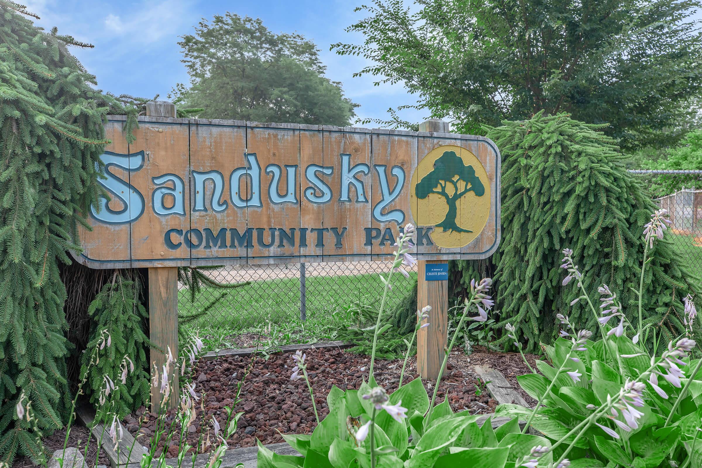 A weathered wooden sign displaying "Sandusky Community Park," surrounded by greenery and flowers. The park's name is prominently featured, with a tree graphic included. The background shows leafed trees and a chain-link fence.