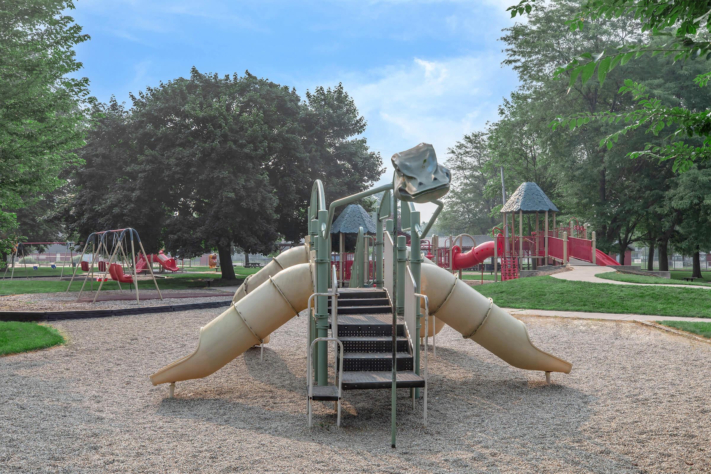 A colorful playground featuring a slide and climbing structures, surrounded by green trees. In the background, additional play equipment like swings and more slides are visible, set on a sandy area under a blue sky.