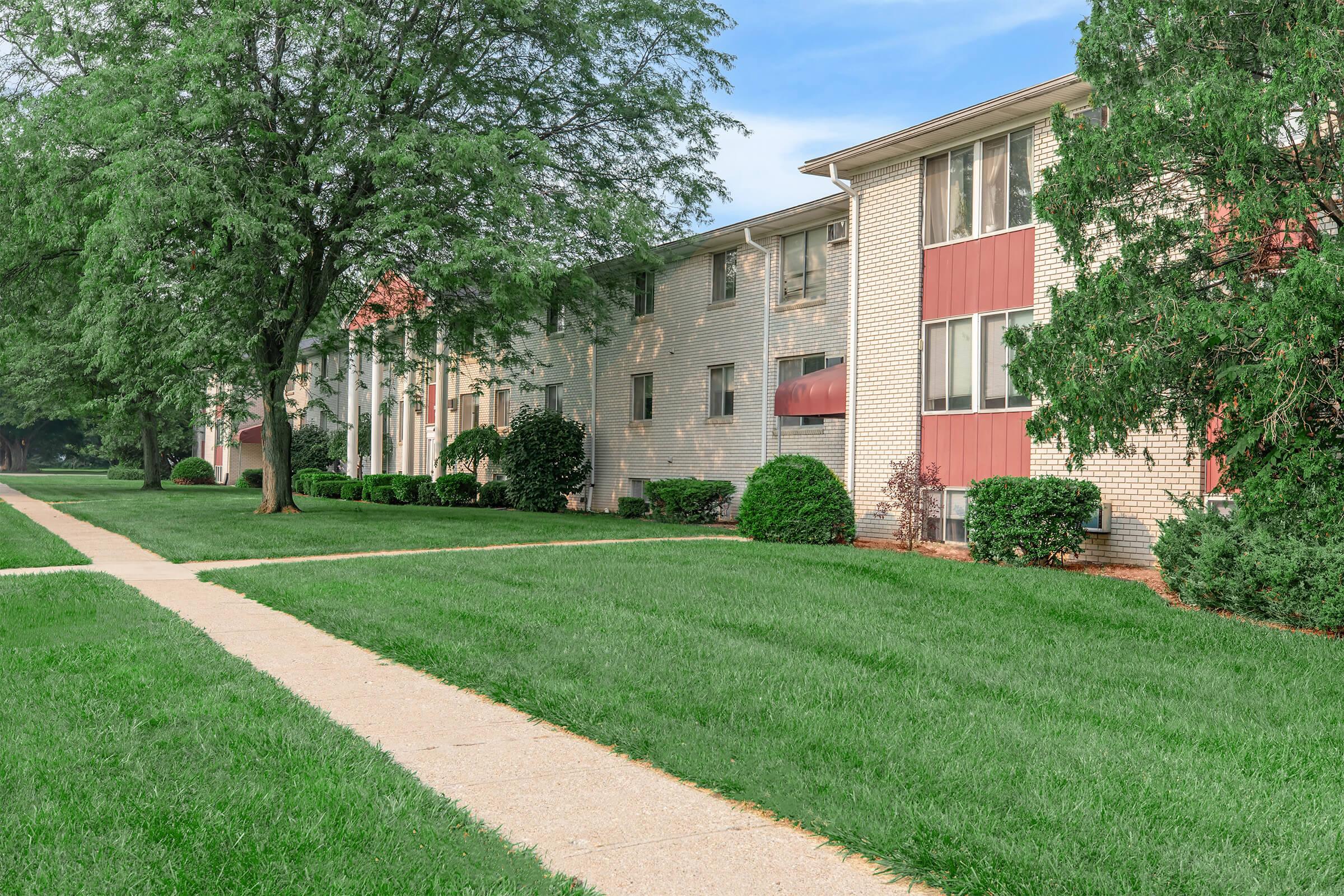 A well-maintained apartment complex with multiple units, featuring red and beige brick exterior. Lush green lawns and neatly trimmed bushes line the front, while a paved walkway leads toward the entrance. Trees provide shade, creating a serene residential atmosphere.