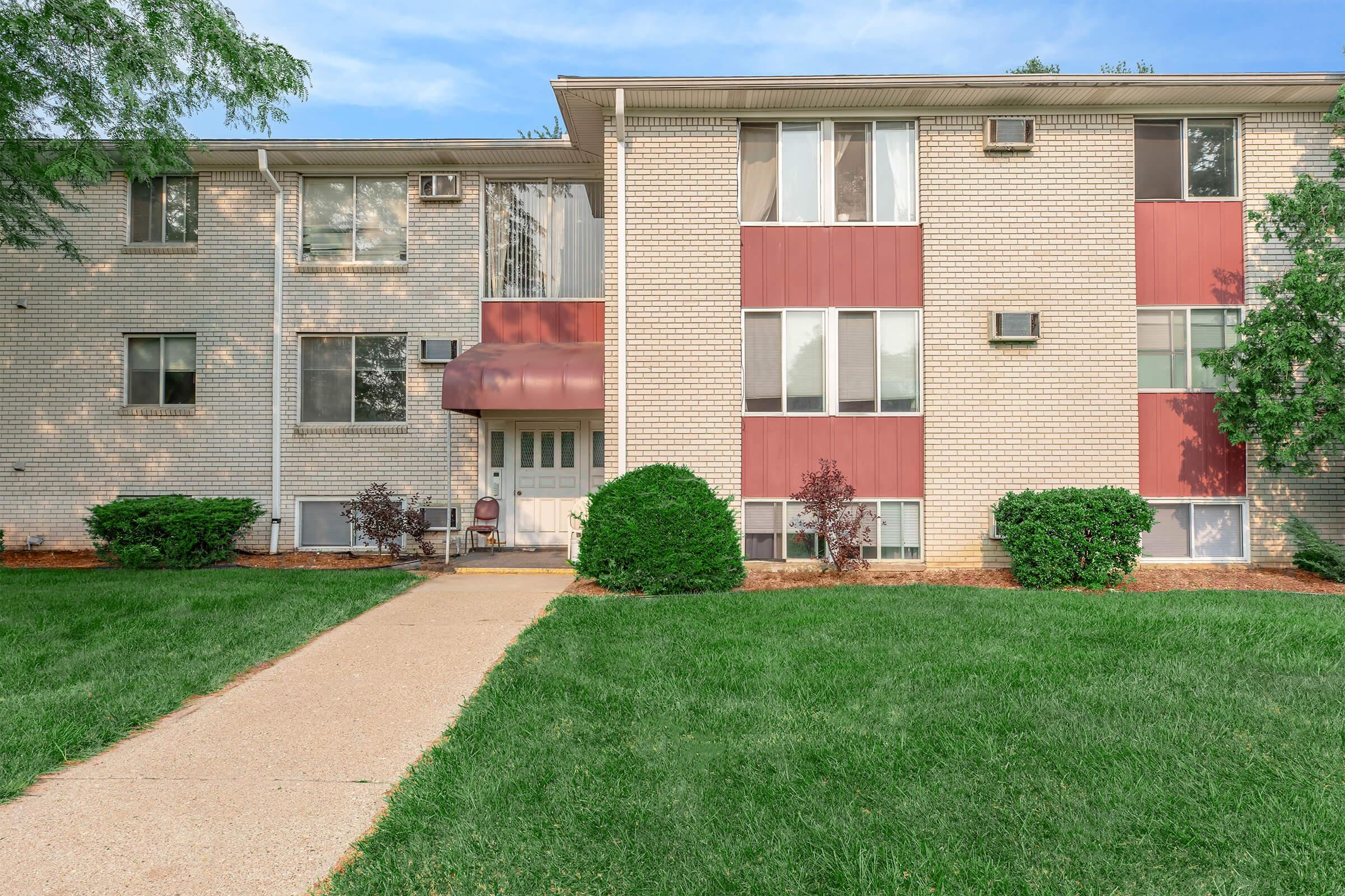 View of a multi-unit residential building with a light-colored brick facade and red accents. The entrance features a covered porch with a chair, surrounded by manicured lawns and small shrubs. A paved pathway leads to the front door, and there are air conditioning units visible on the exterior walls.
