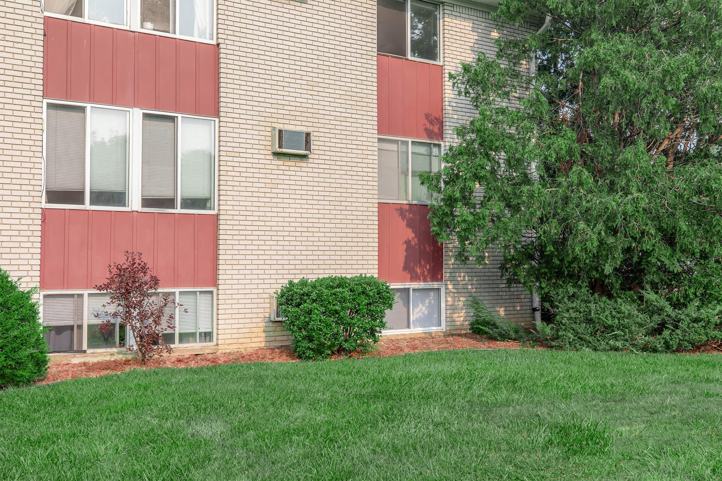 A low-rise brick apartment building featuring large windows with red accents. There are small shrubs and grass in the foreground, and a tree to the right, providing a green landscape around the building. An air conditioning unit is visible on the side of the building.