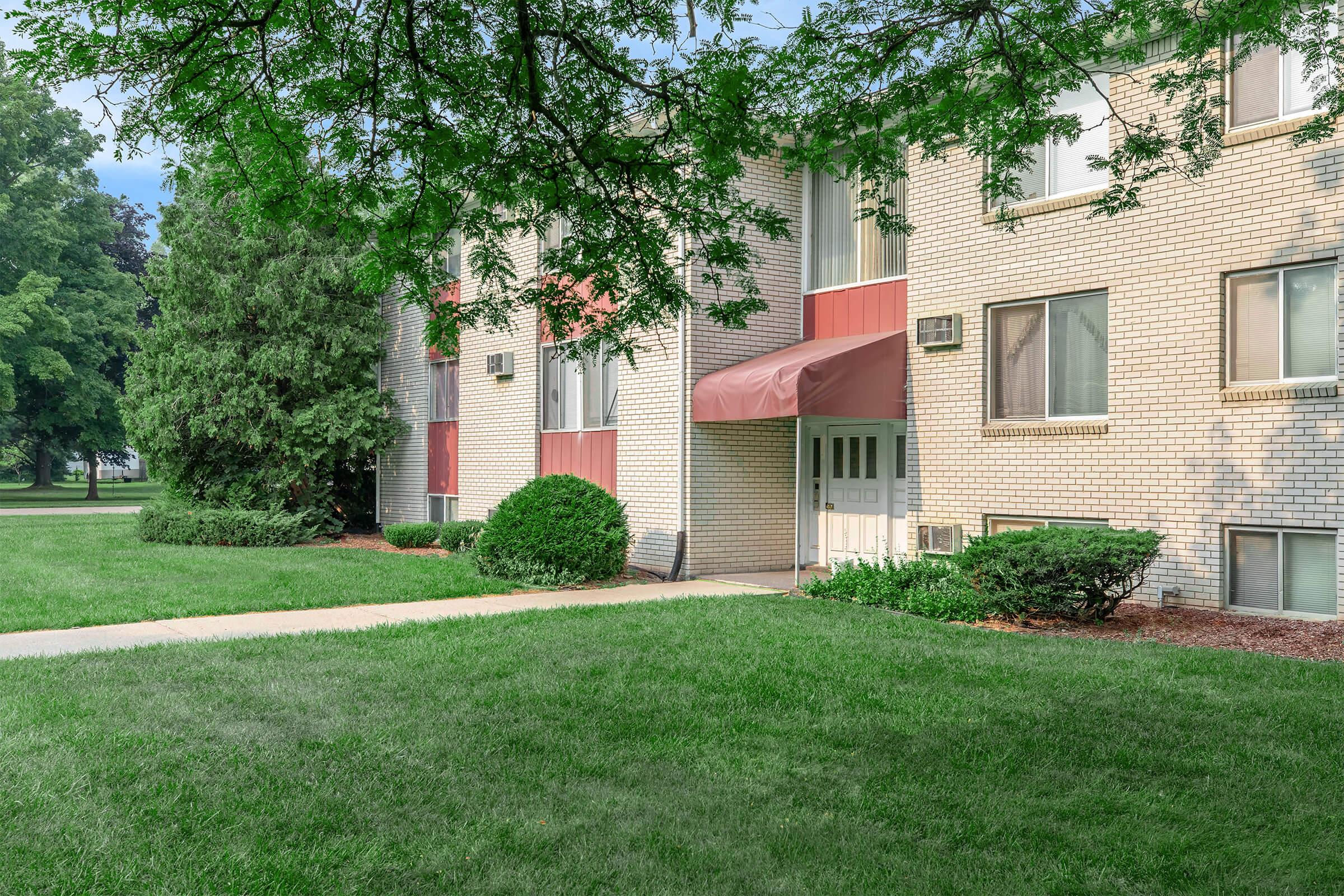 Exterior view of a brick building with large windows. There is an entrance with a maroon awning and a landscaped area featuring green grass, shrubs, and trees. The setting is well-maintained and surrounded by a clear sky.