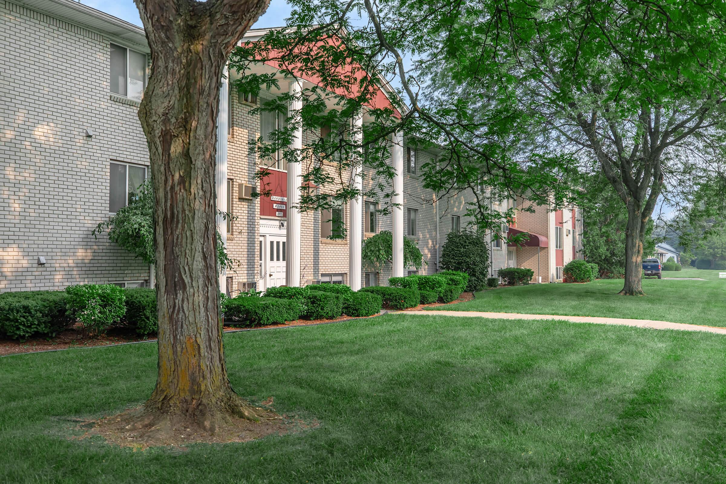 Exterior view of a residential building with brick facade, featuring columns and a welcoming entrance. The scene includes a well-maintained lawn with a tree in the foreground and shrubs lining the pathway. Soft sunlight filters through the leaves, creating a peaceful atmosphere.