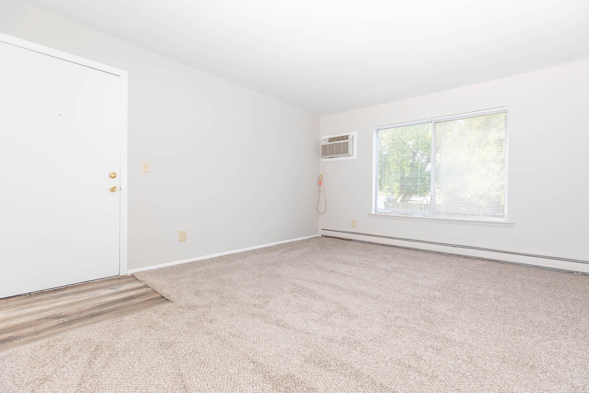 A light-filled interior of a room with beige carpet, white walls, and a large window. An air conditioning unit is installed beneath the window, and there is a simple white door on the left. The space appears empty and ready for decorating or furniture placement.