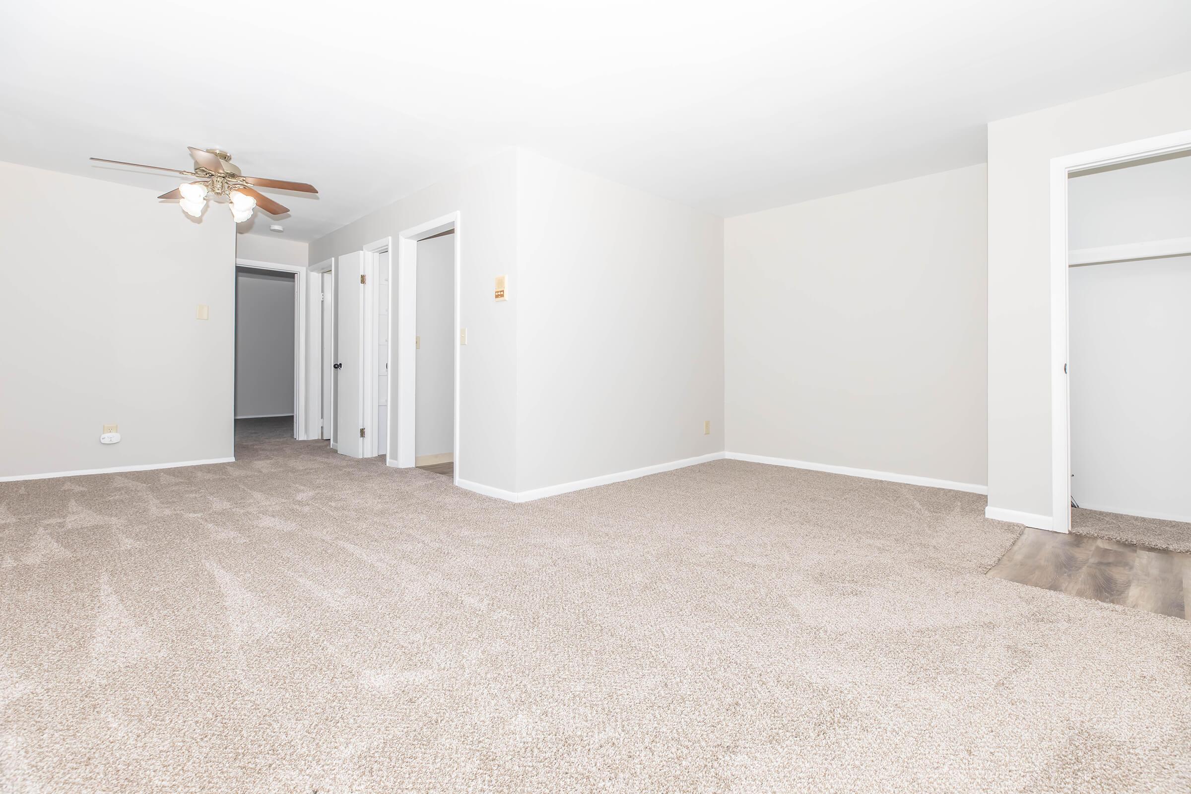 A spacious, well-lit interior of a room featuring light gray walls, beige carpet flooring, and a ceiling fan. Multiple doorways lead to adjacent rooms, with one corner showing a partial wall and closet space. The overall ambiance is clean and inviting, suitable for various furniture arrangements.