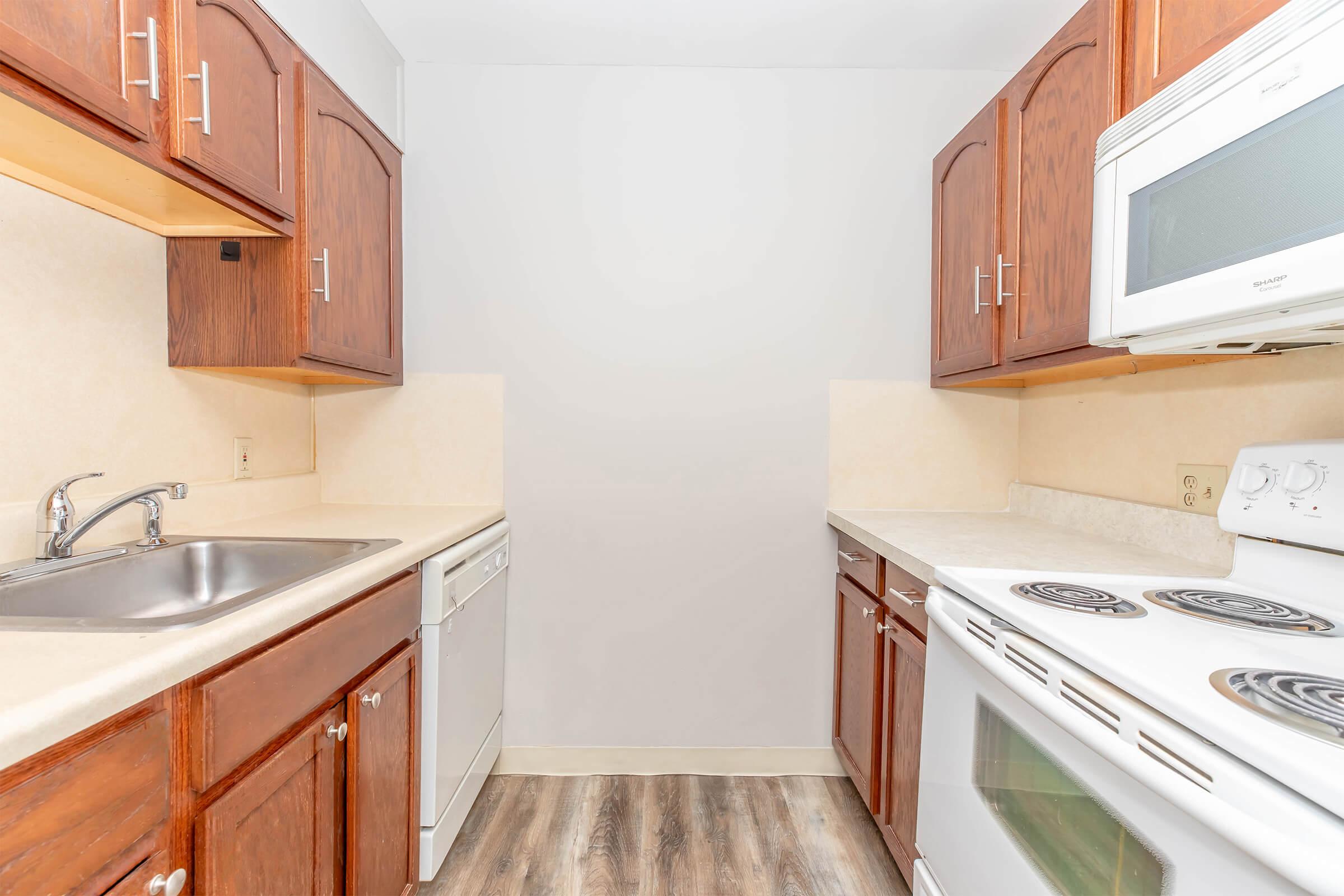 A compact kitchen featuring wooden cabinets, a stainless steel sink, a dishwasher, and a white stove with an oven. The walls are light-colored, and the flooring is a wood-like laminate. Natural light brightens the space, creating a clean and inviting atmosphere.