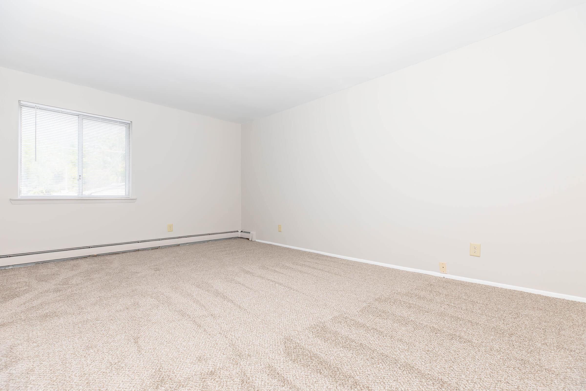 An empty room with light beige walls and a window allowing natural light. The floor is covered with light-colored carpet, and there are minimal furnishings. The space appears clean and ready for decoration or furnishing.