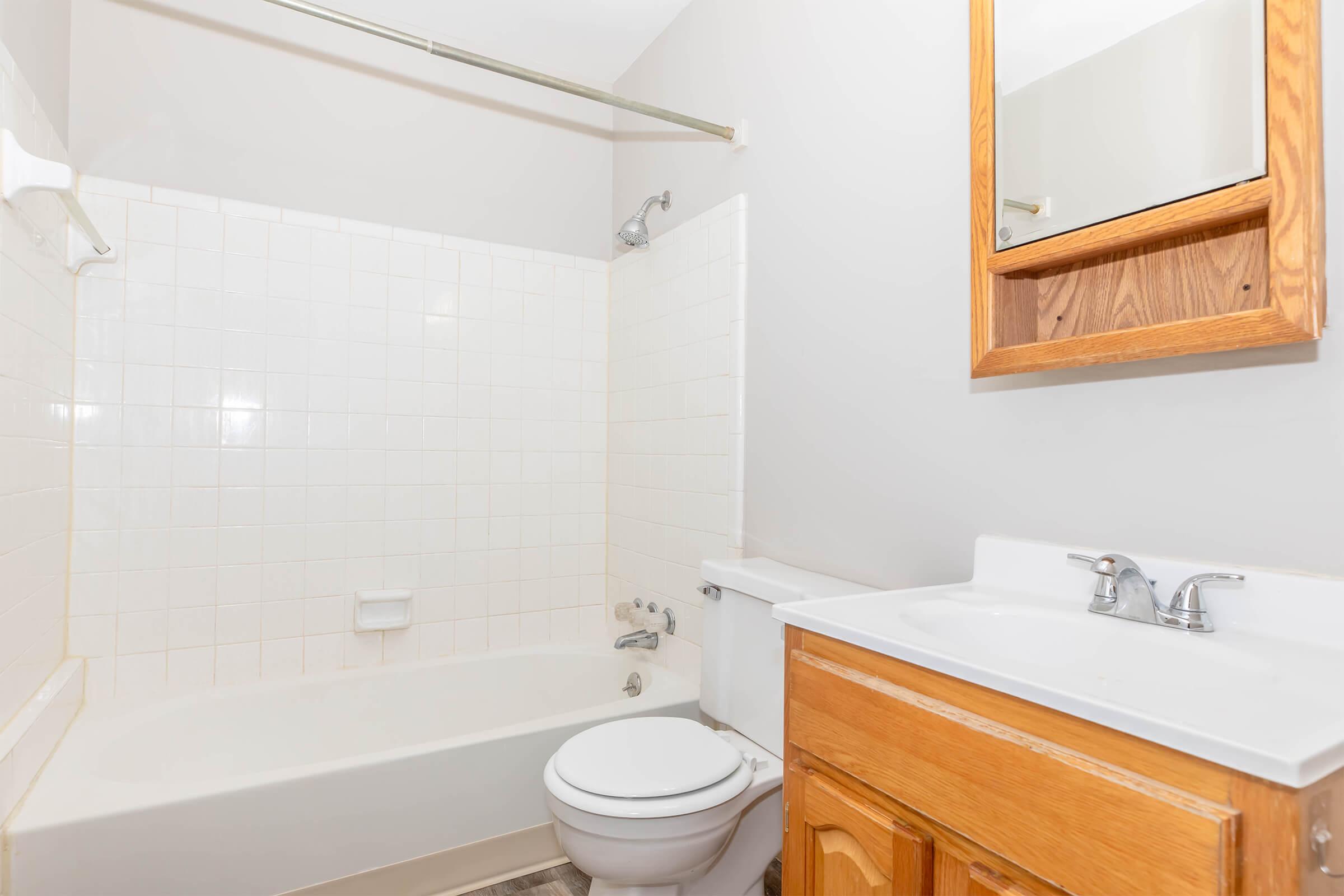 A clean, simple bathroom featuring a white bathtub with a showerhead, a toilet, and a wooden vanity with a sink. The walls are painted light gray, and a mirror is mounted above the sink. The overall design is minimalist and functional, with tiled walls in the shower area.