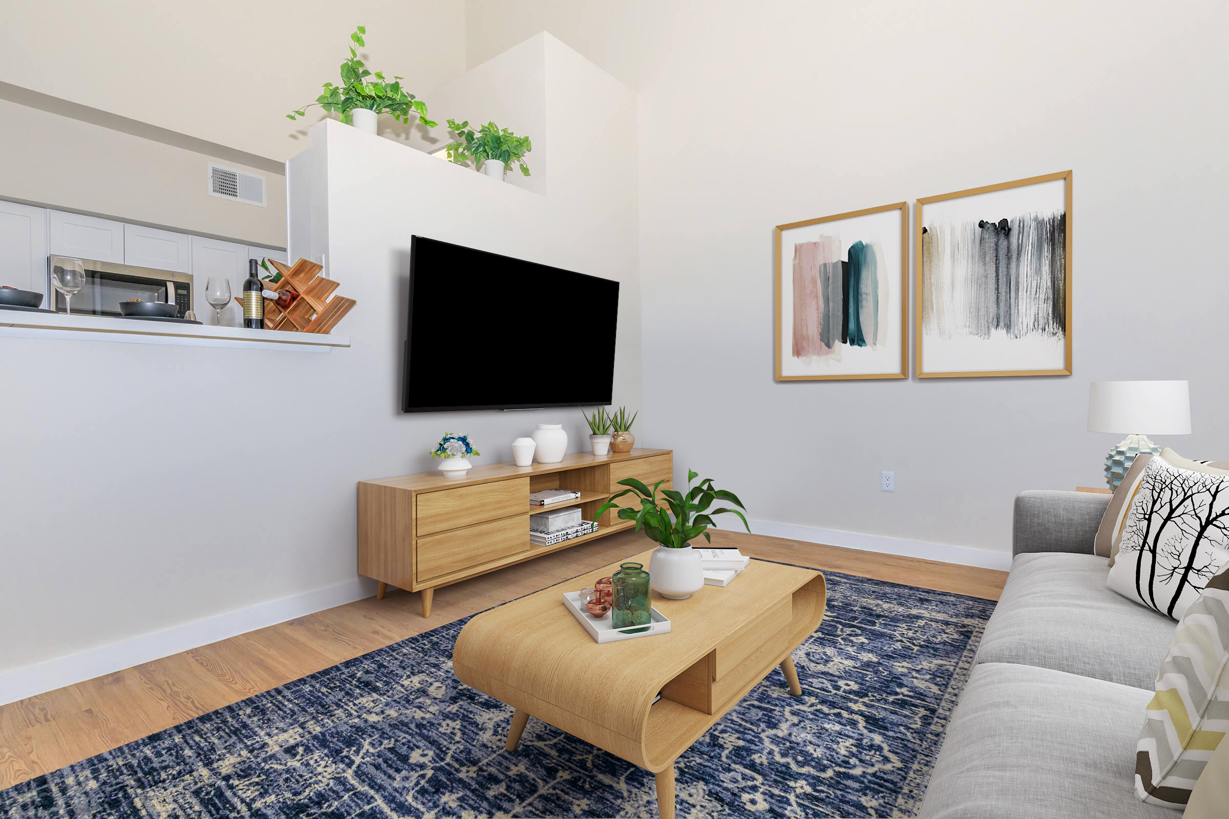 A modern living room featuring a light color scheme, a gray sofa, and a wooden coffee table. There is a large TV mounted on the wall, with decorative items on a media console beneath it. Two framed artworks hang above the console, and a plant adds greenery to the space. The floor is wooden, with a patterned area rug.