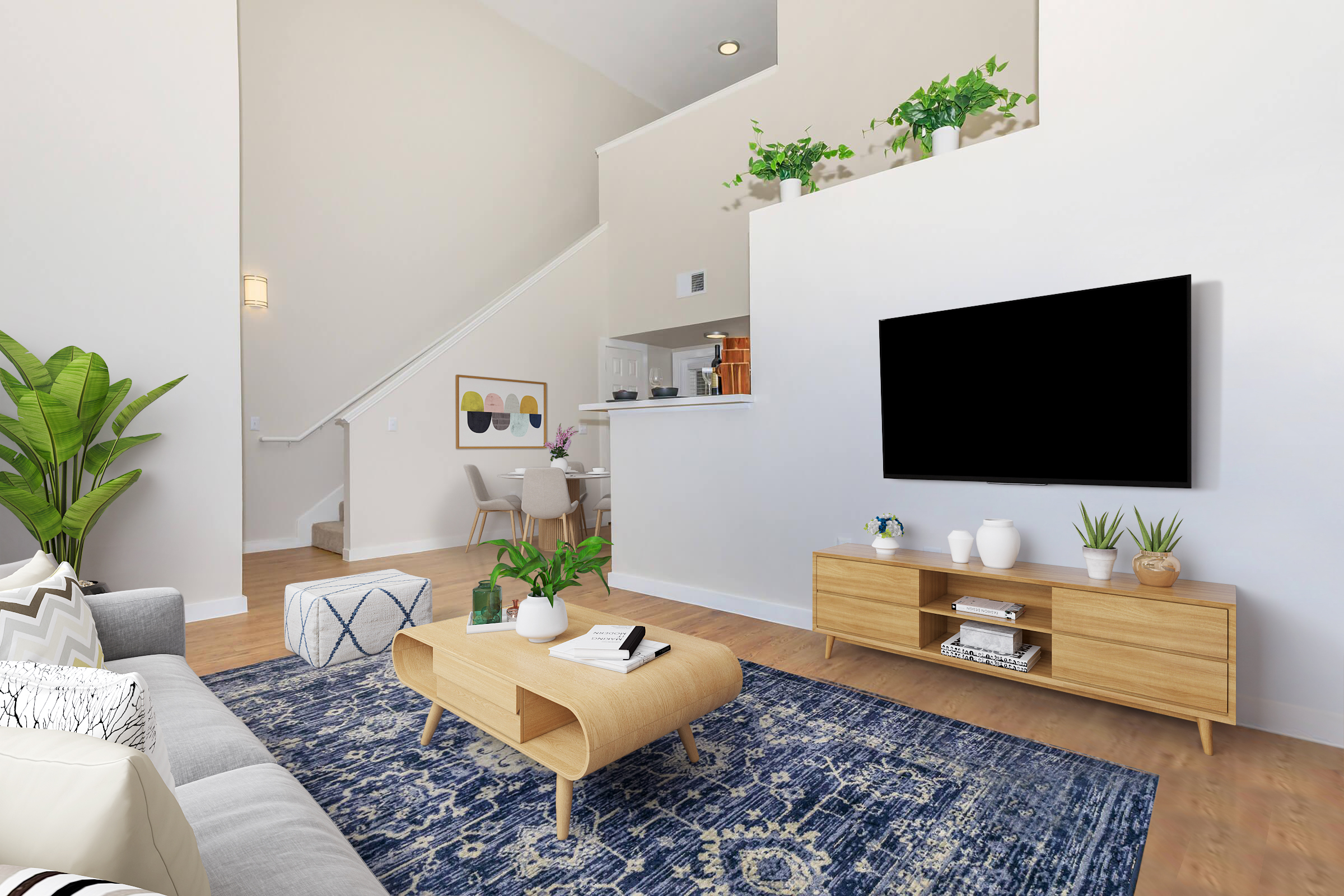 A modern living room featuring a light-colored sofa with decorative pillows, a wooden coffee table, and a large flat-screen TV on a light wood media console. The space includes a blue area rug and potted plants, with a dining area visible in the background. Natural light fills the room, creating a welcoming atmosphere.