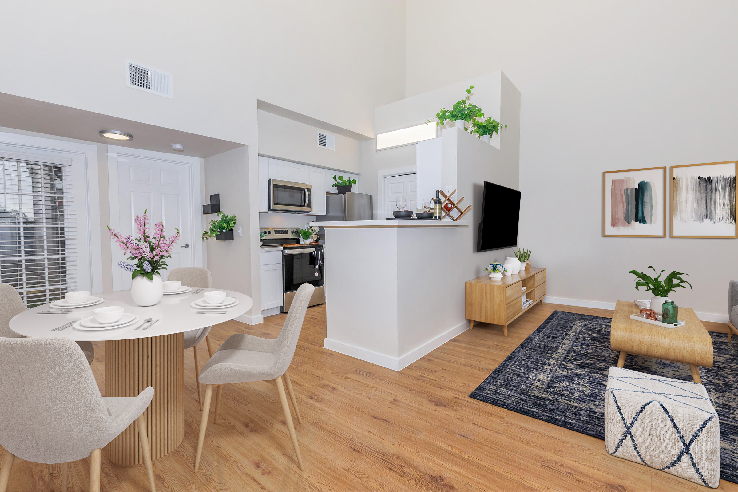 A modern open-concept living space featuring a dining area with a round table set for four, a sleek kitchen with stainless steel appliances, and a cozy living room with a television and decorative plants. The decor includes light wood finishes, a large area rug, and framed artwork on the walls.